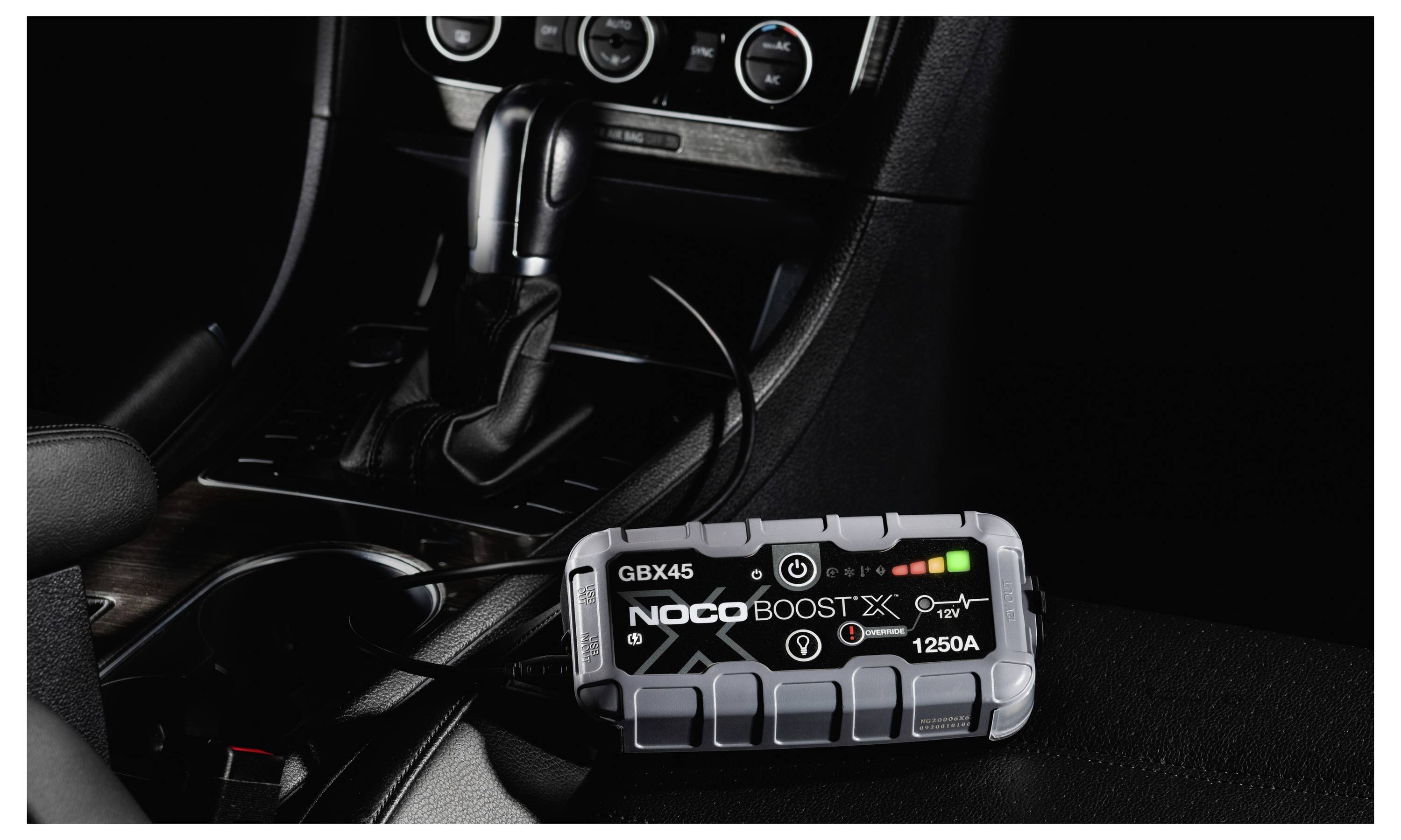 Přenosná startovací souprava NOCO Boost X 1250A leží na sedadle automobilu vedle středové konzole a řadící páky s připojeným kabelem.