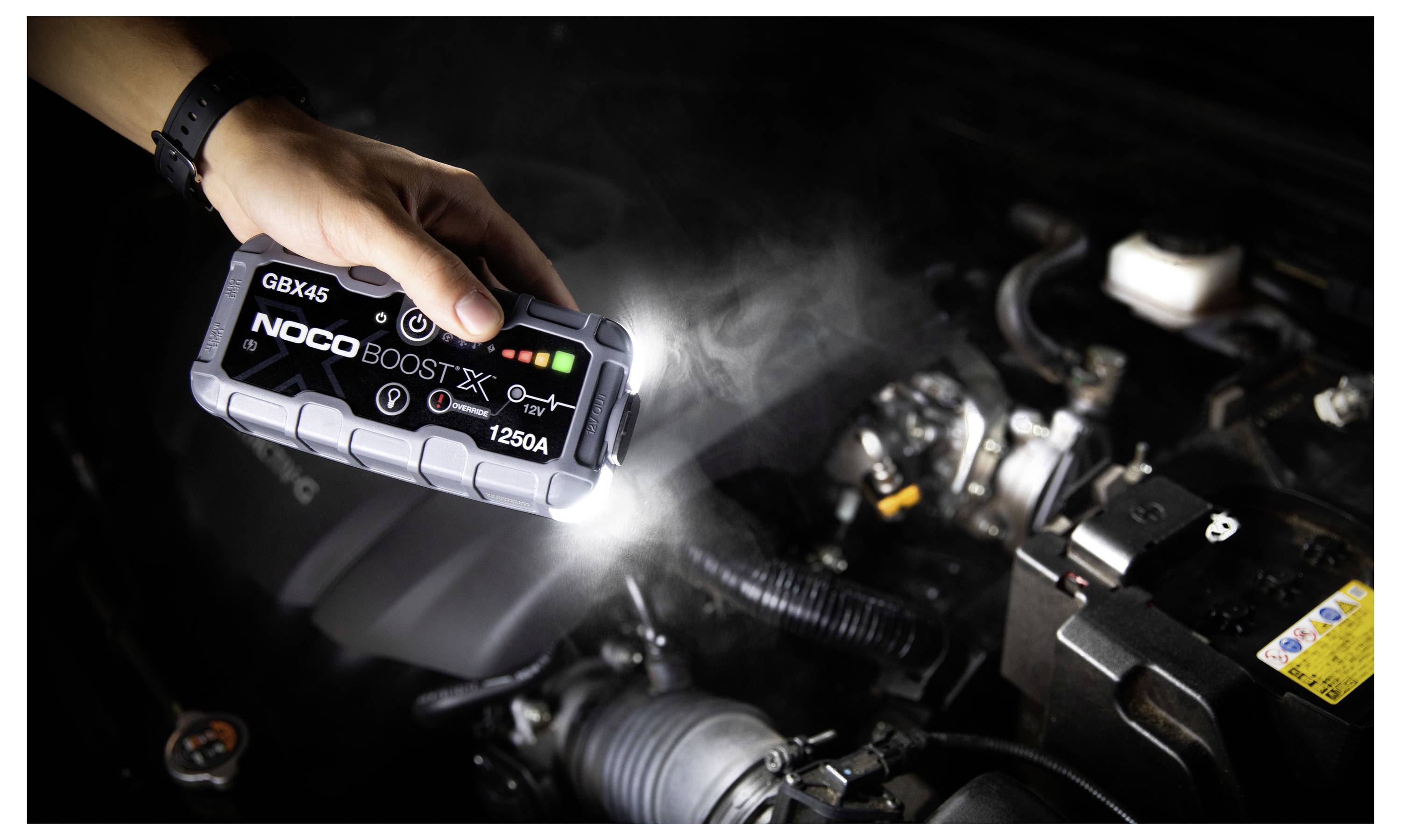 Ruka drží přenosný startér NOCO Boost X GBX45 v motorovém prostoru automobilu a používá jeho svítilnu k kontrole součástek.
