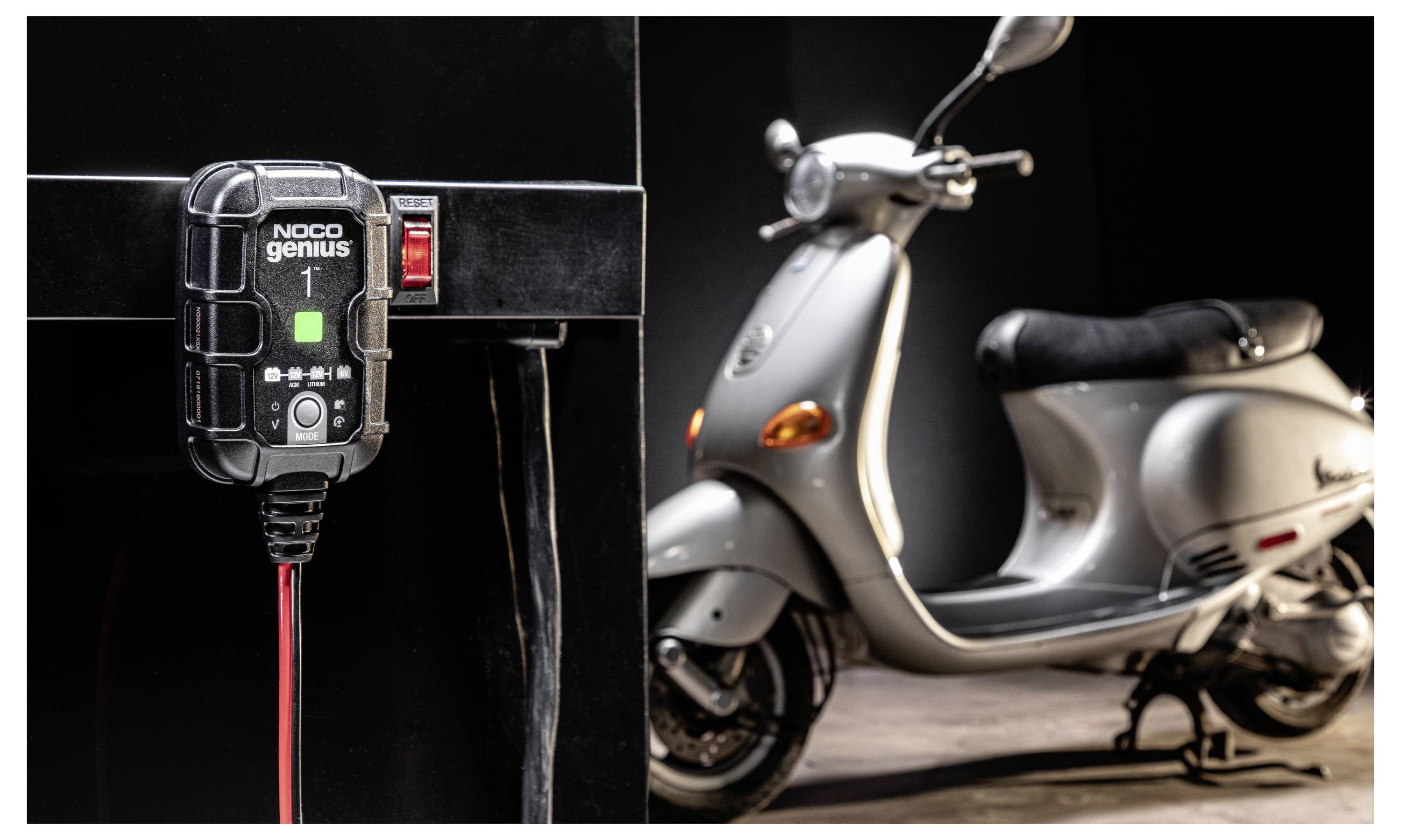 Nabíječ baterií NOCO Genius je zastrčen do nabíjecího bodu elektrokola v garáži, přičemž elektrokolo je zaparkováno v pozadí.