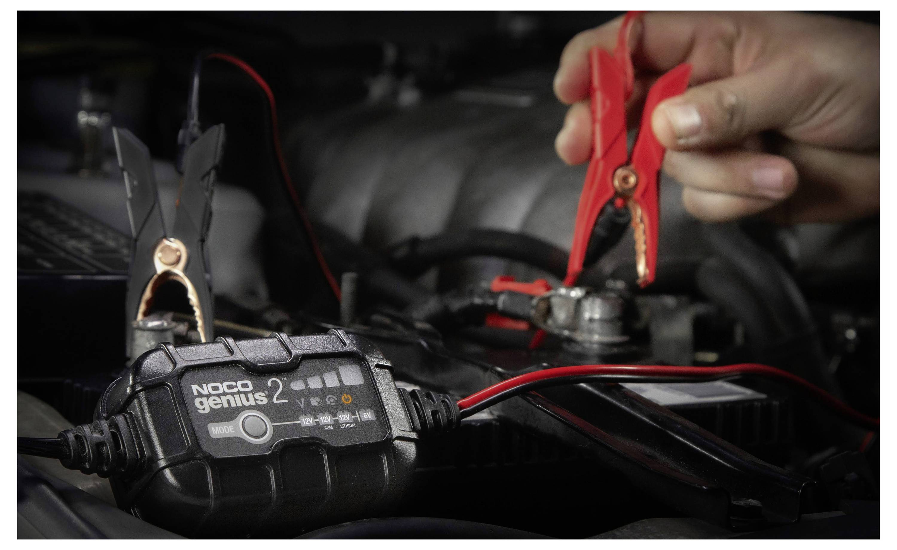 Ruce používající červené a černé svěrky k připojení nabíječky baterií NOCO Genius k autobaterii v motorovém prostoru.