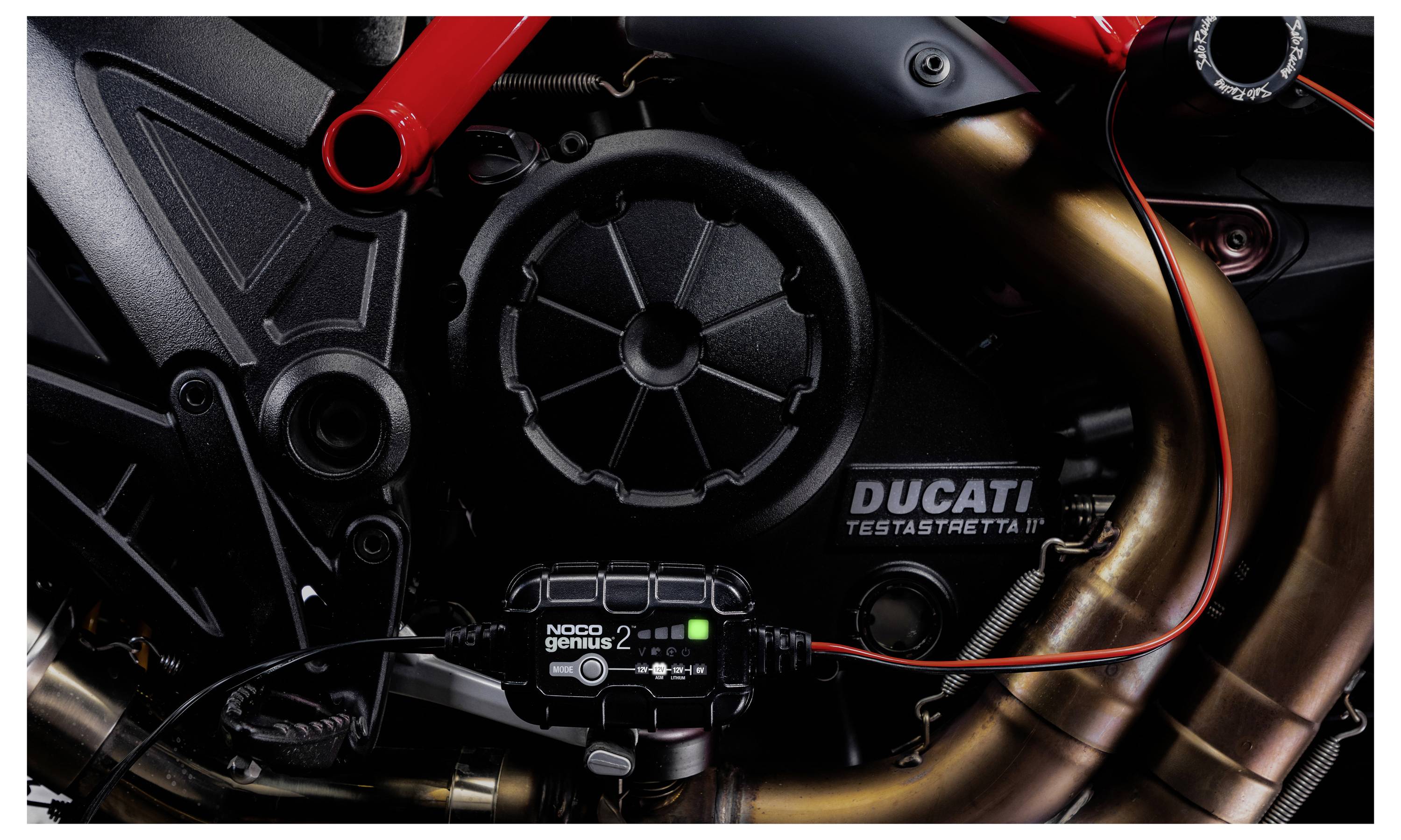 Detailní pohled na motor motocyklu Ducati s brandingem "Ducati Testastretta" a připojeným elektronickým modulem, zobrazující červený rám a výfukové potrubí.