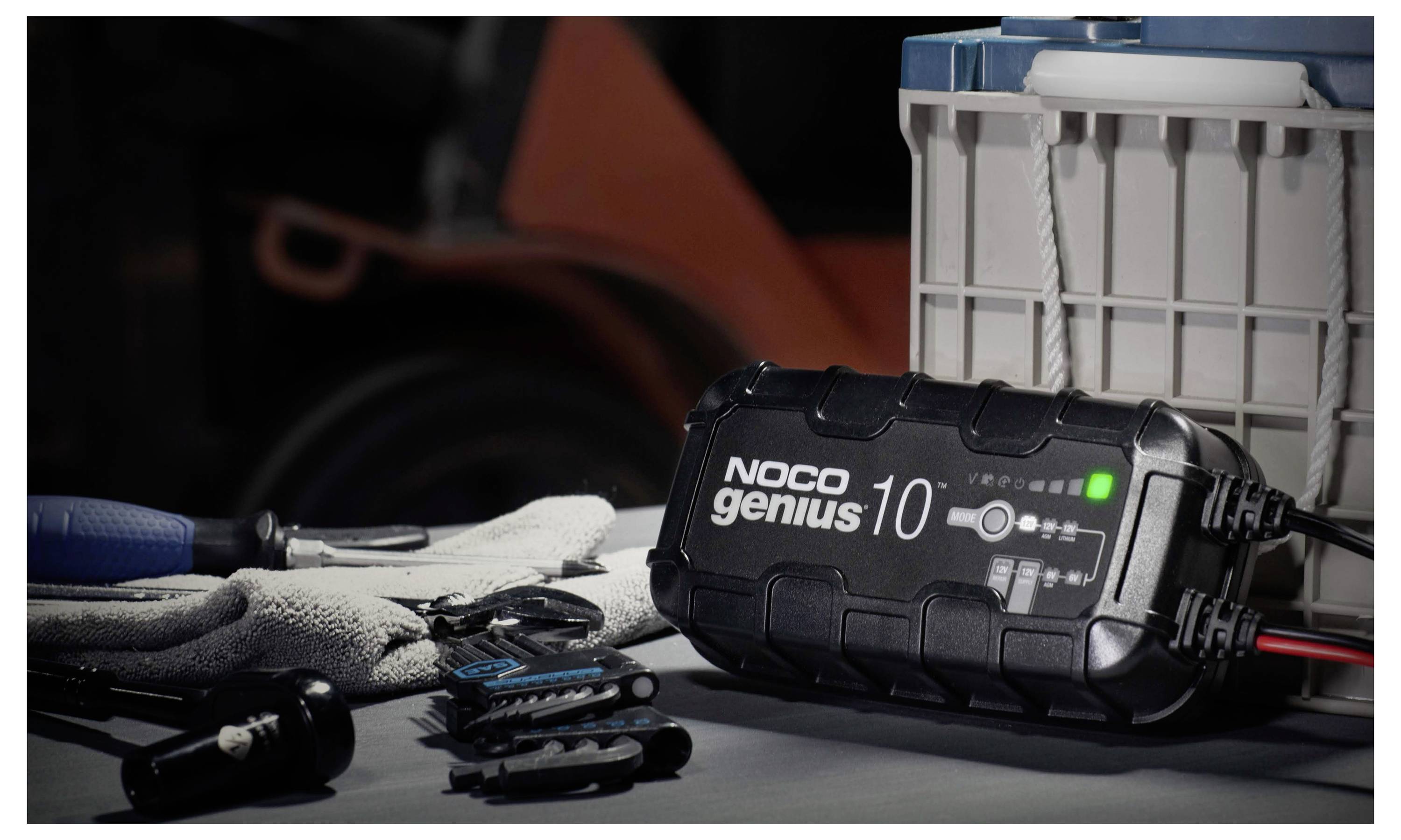 Nabíječ baterií NOCO Genius 10 stojí na pracovním stole vedle pracovních rukavic, ručních nástrojů a plastového skladovacího boxu.