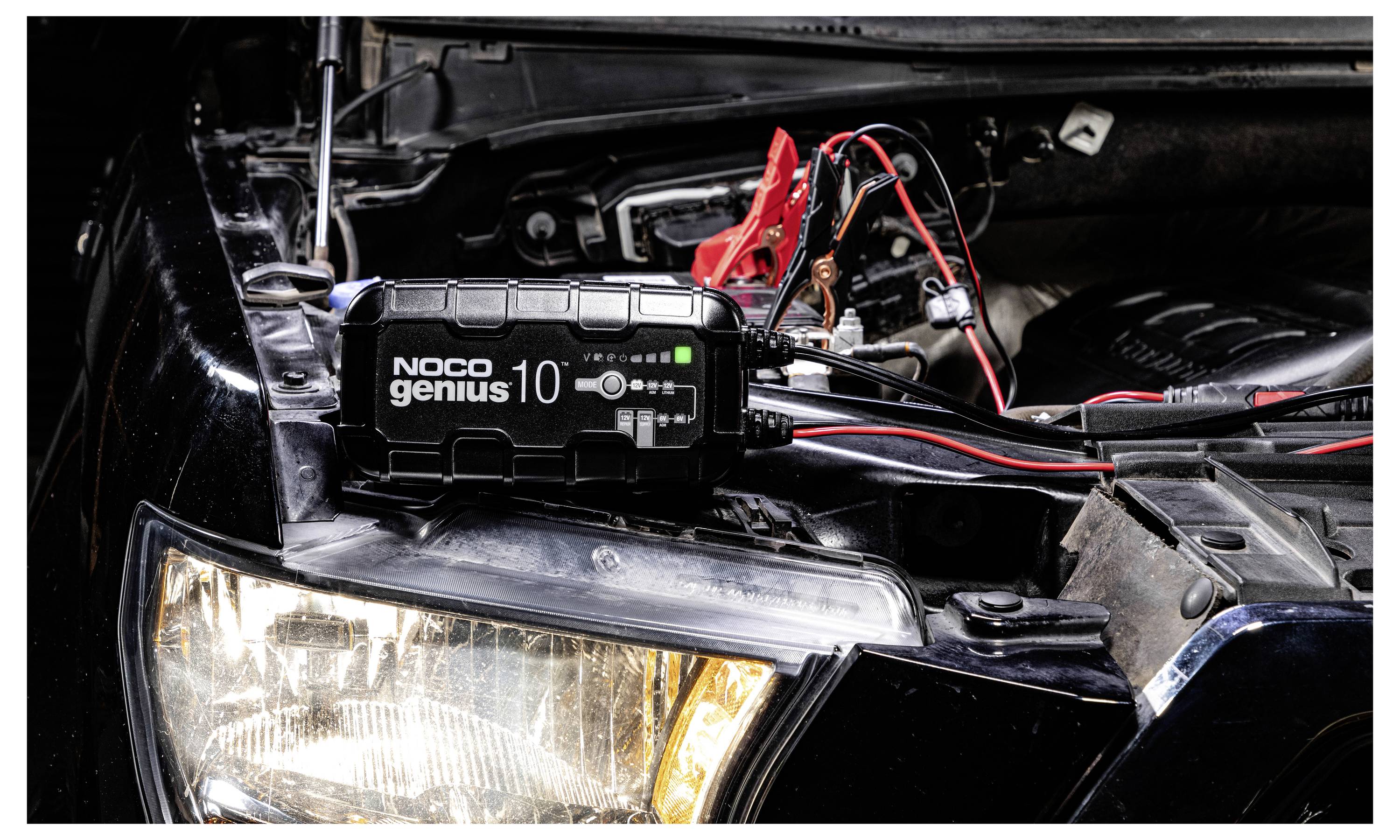 Přenosná nabíječka baterie NOCO Genius 10 připojená k autobaterii v otevřeném motoru, s připojenými červenými a černými svorkami.