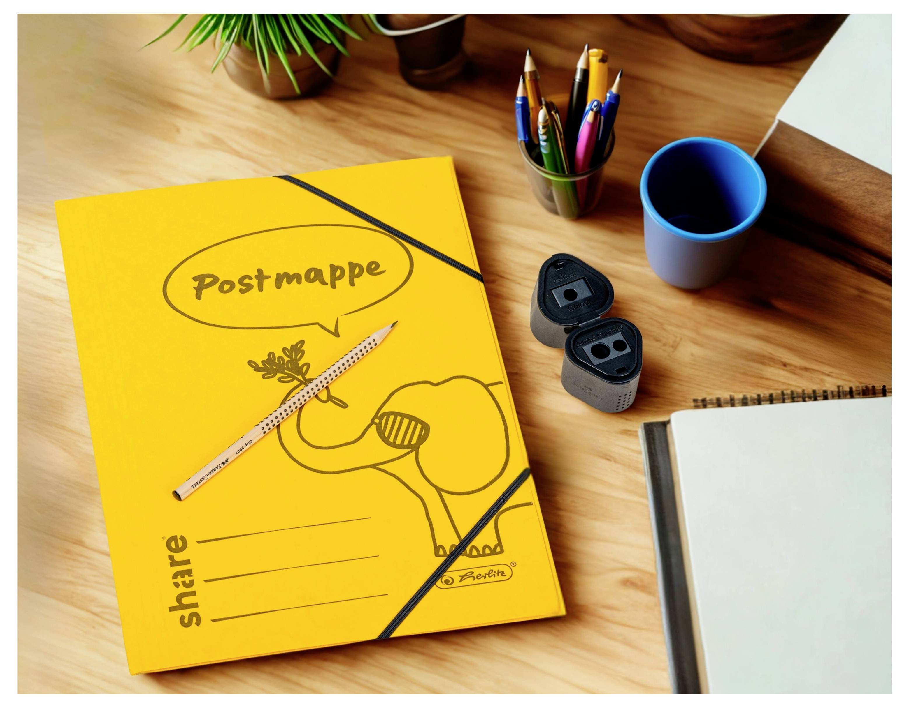 Žlutá složka "Postmappe" na stole s tužkami, šálky a poznámkovým blokem, sugerující kancelářský pracovní prostor nebo pracovní plochu.