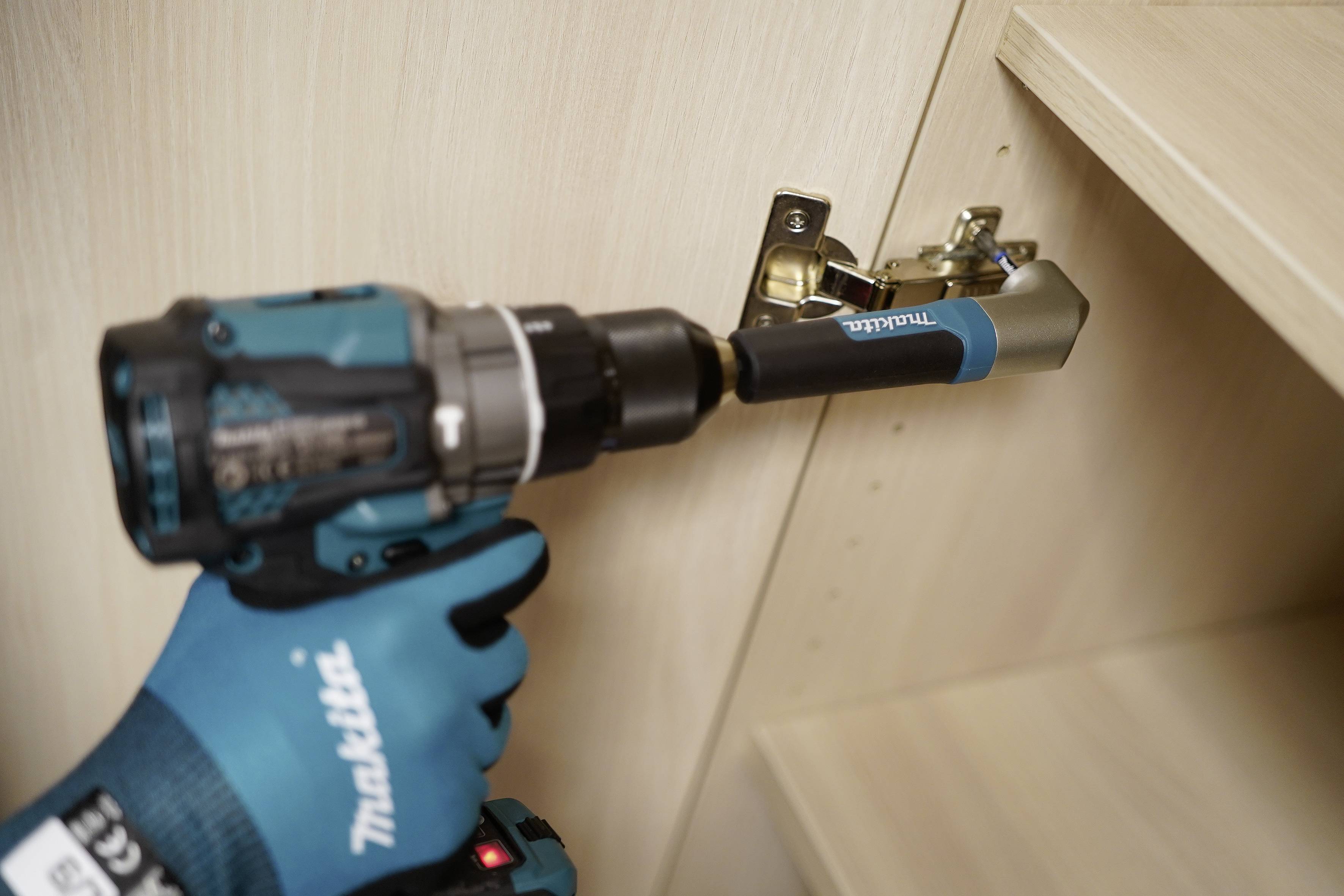 Rukavicí oděná ruka používá bezdrátový vrtací přístroj Makita k instalaci pantázu skříně uvnitř dřevěné skříně.