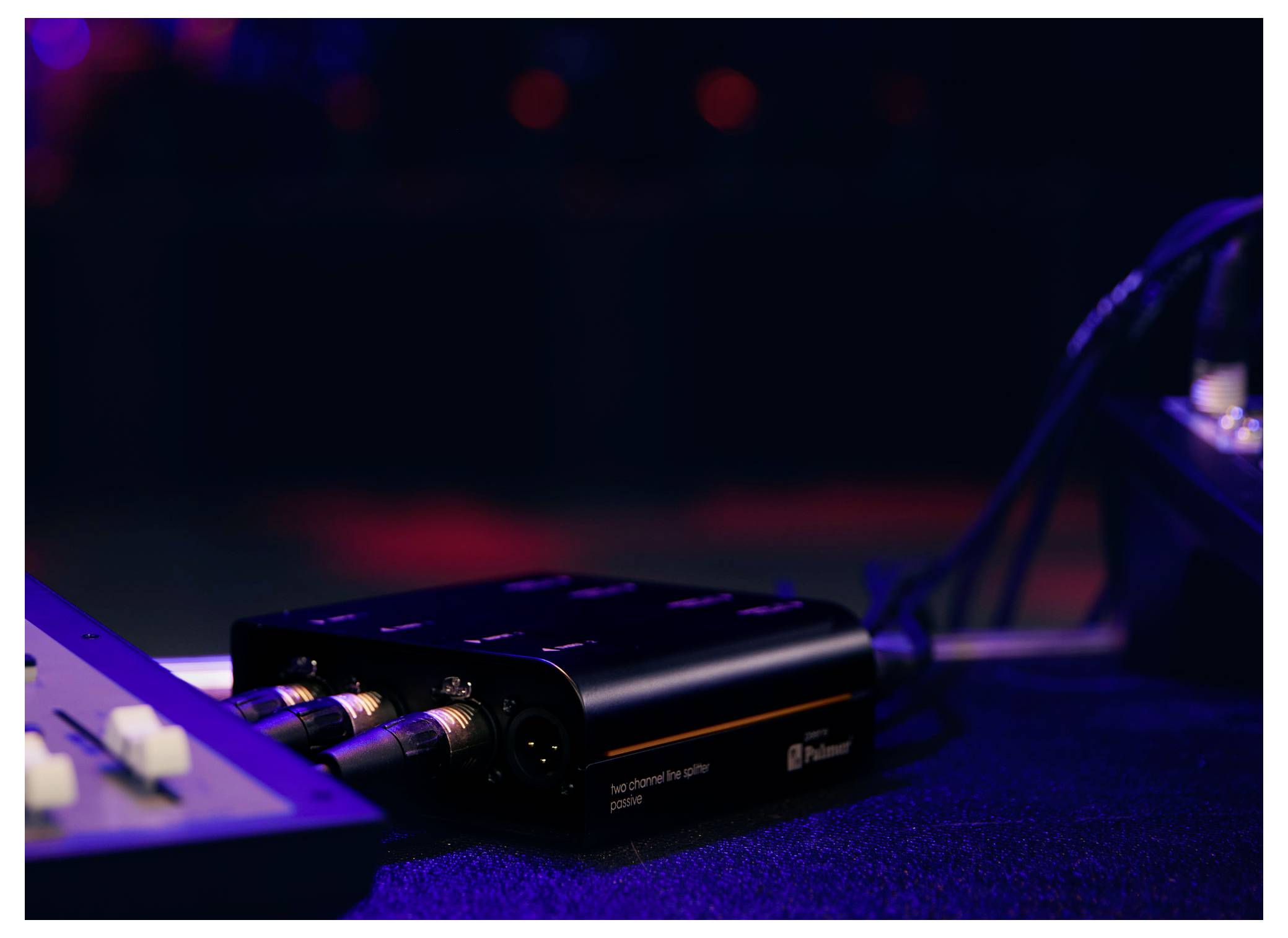 Detailní záběr černého audio rozhraní nebo adaptéru na stole s připojenými kabely a rozostřeným osvětlením podobným osvětlení na scéně v pozadí.
