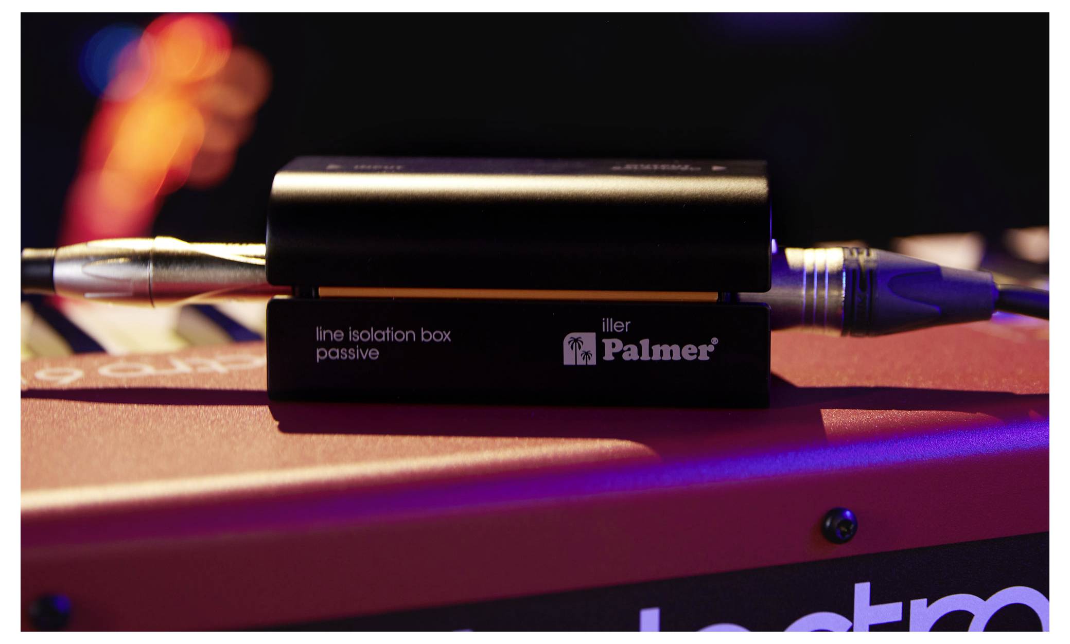Detailní pohled na pasivní izolační box Palmer s audio kabely zapojenými na obou koncích, spočívajícím na červeném povrchu pod scénickým osvětlením.