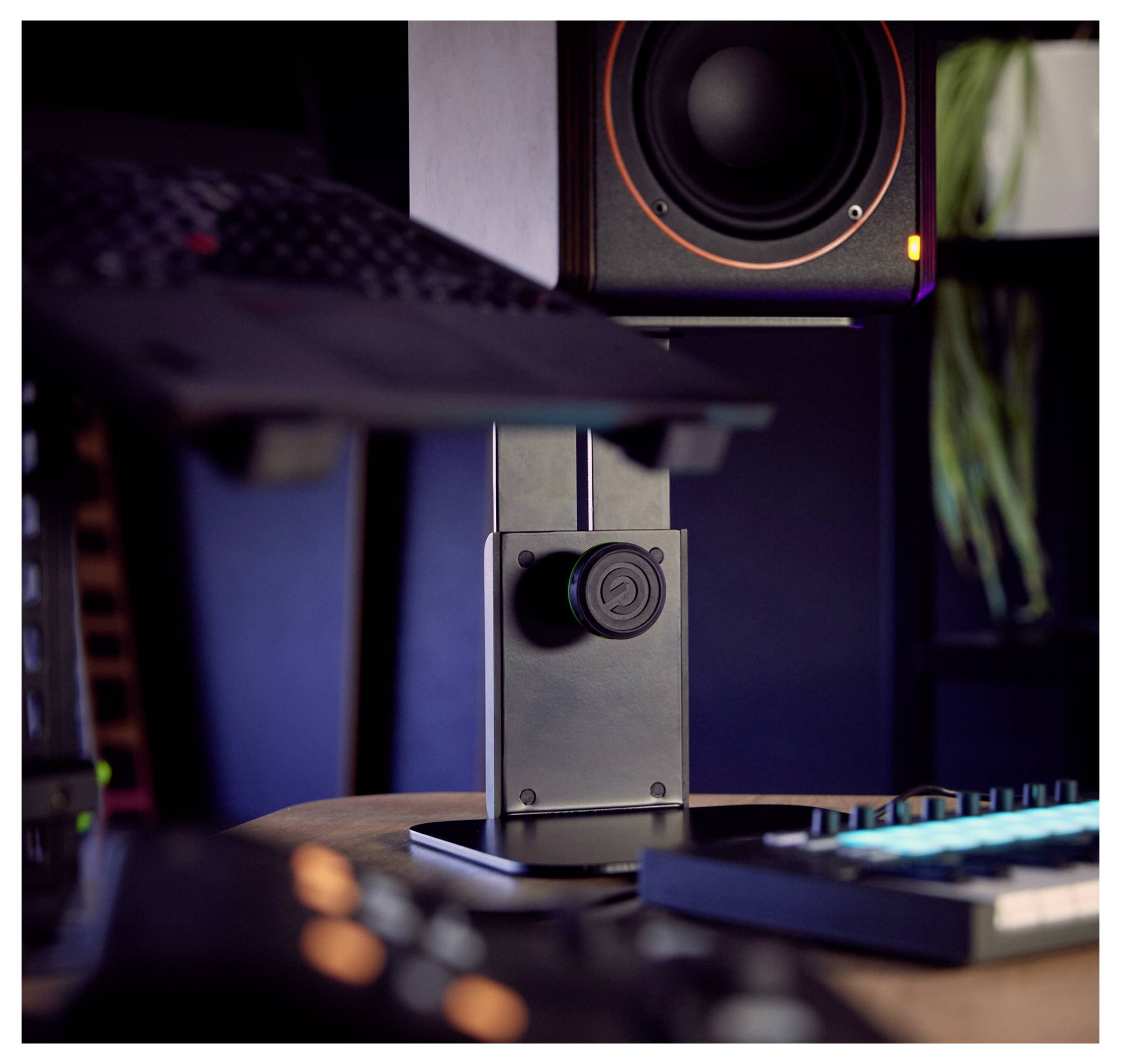 Detailní záběr černého stolního stojanu pro reproduktor s zvukovým ovladačem, vedle klávesnice a monitoru v tmavém studiovém nastavení.