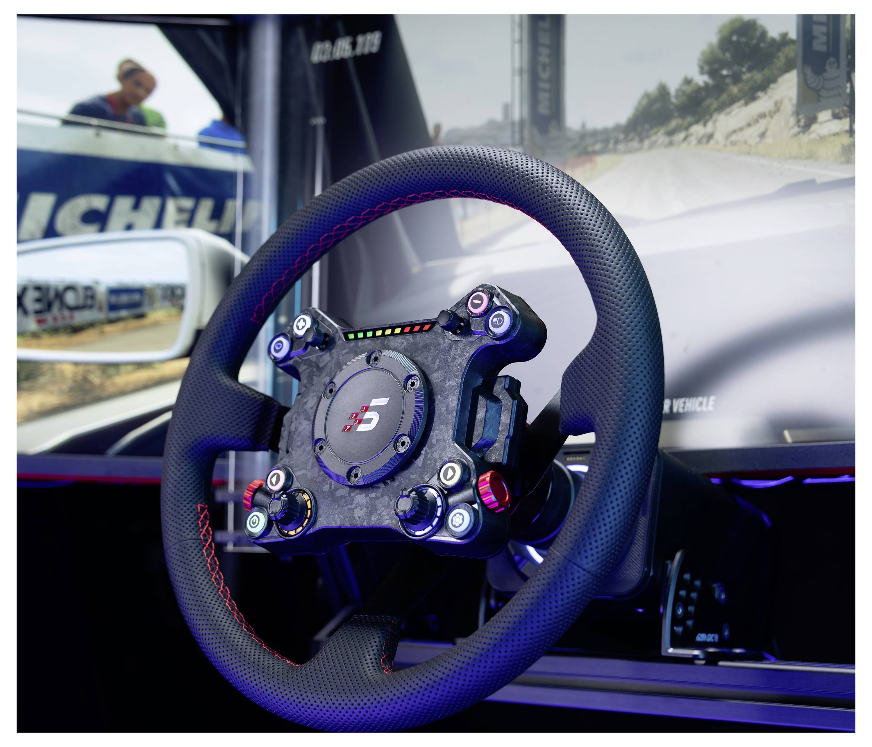 Detailní záběr na volant závodního vozu namontovaný v simulátoru nebo kokpitu pro předvádění, s rozmazanou závodní scénou a brandingem Michelin v pozadí.