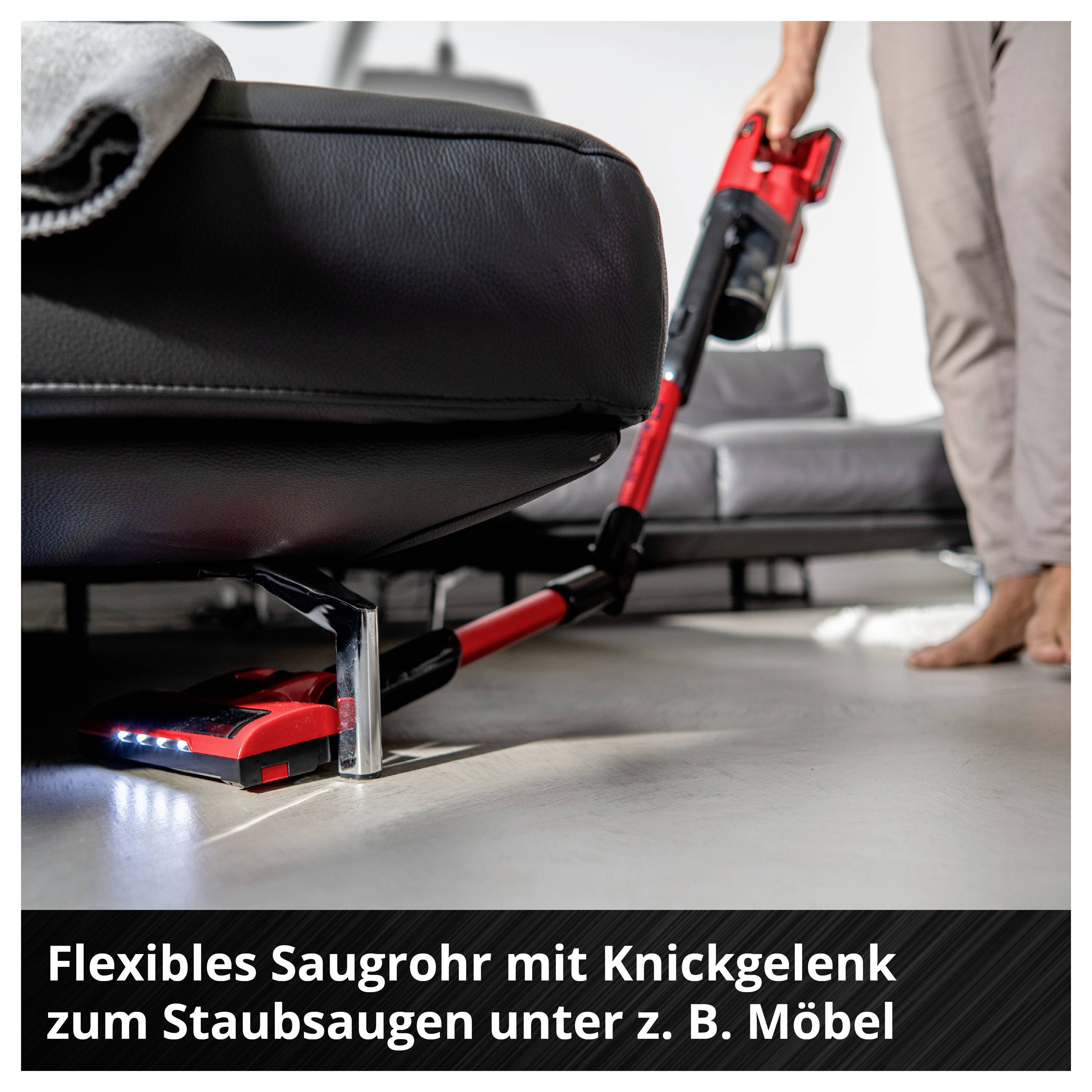 Červený akumulátorový vysavač s kloubem čistí pod gaučem v obývacím pokoji; Poznámka: Flexibilní sací hadice pro vysávání pod nábytek.