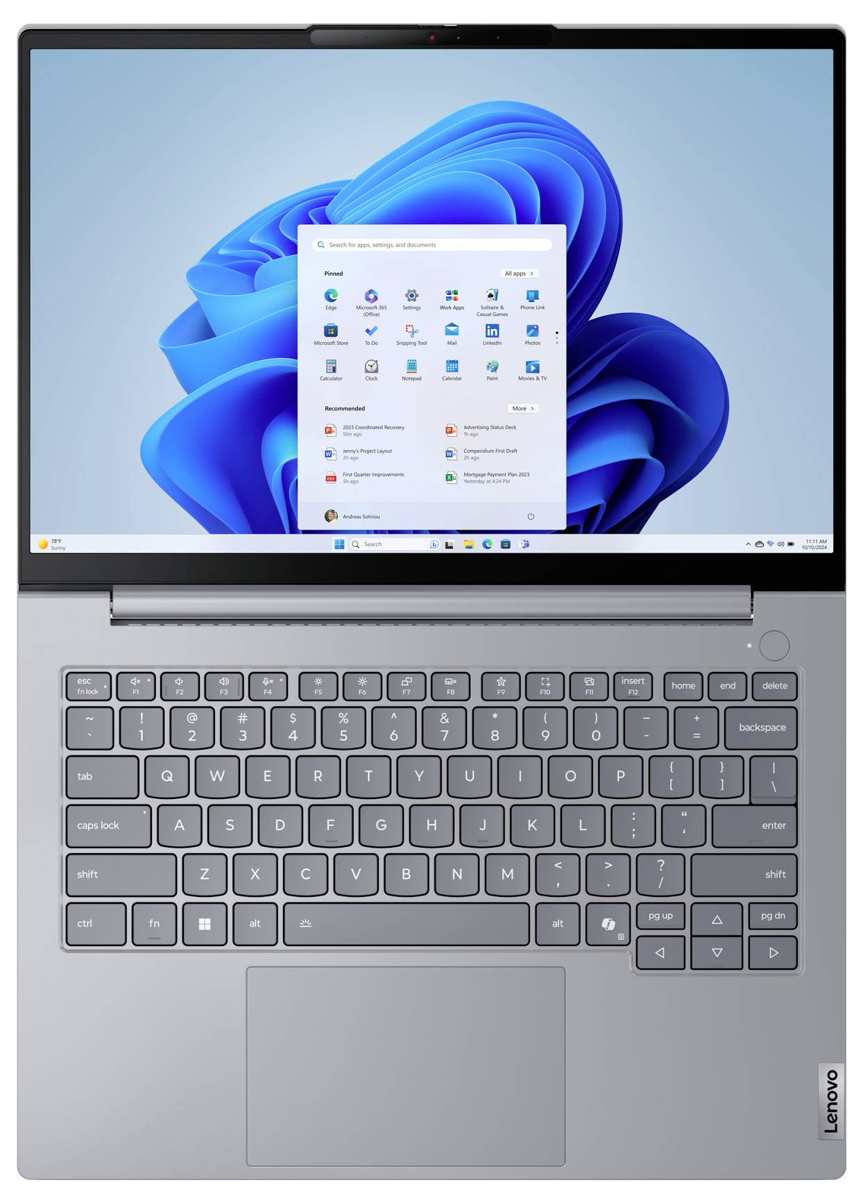 Šedý notebook Lenovo se systémem Windows 11 otevřený na ploše, zobrazující středově umístěnou nabídku Start na modrém abstraktním pozadí.