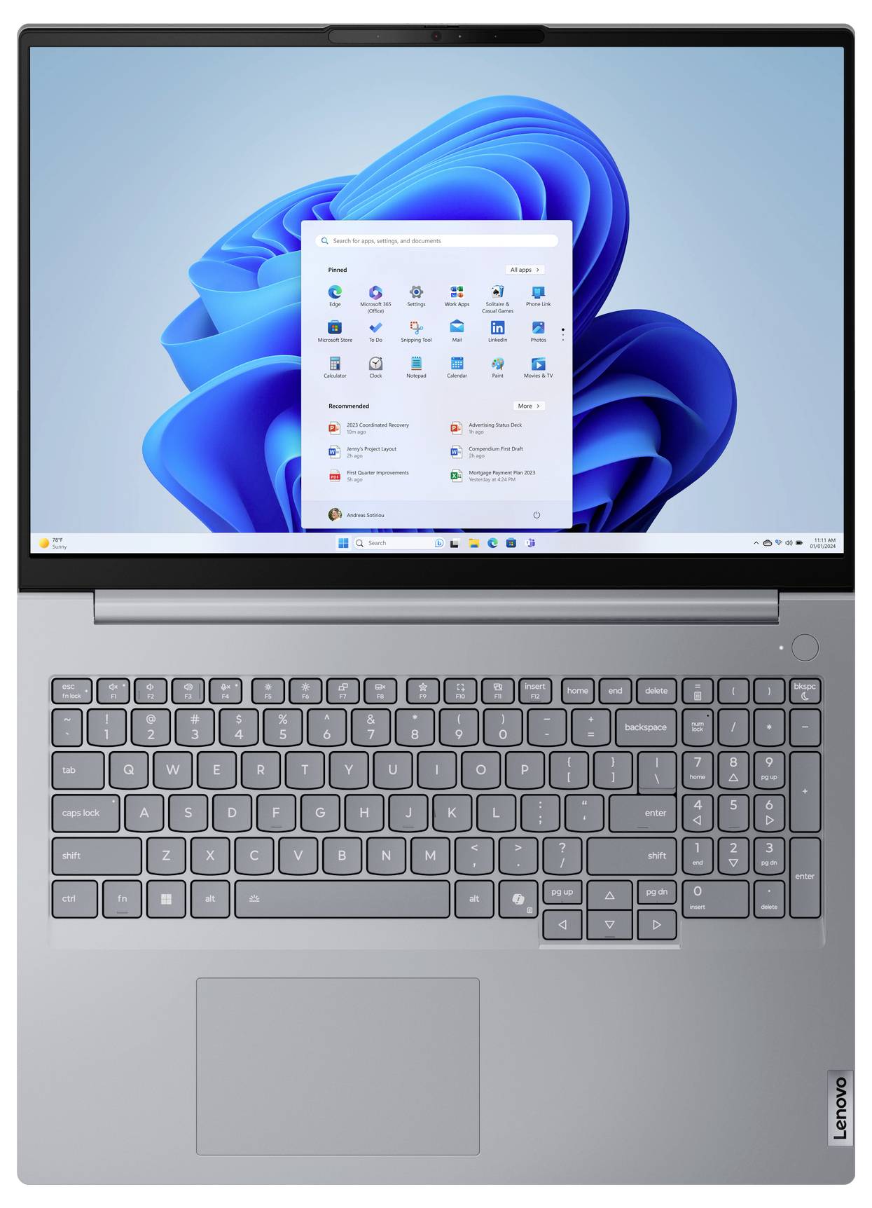 Stříbrný laptop Lenovo otevřený se stolem Windows 11 a nabídkou Start na obrazovce, zobrazující klávesnici a touchpad.