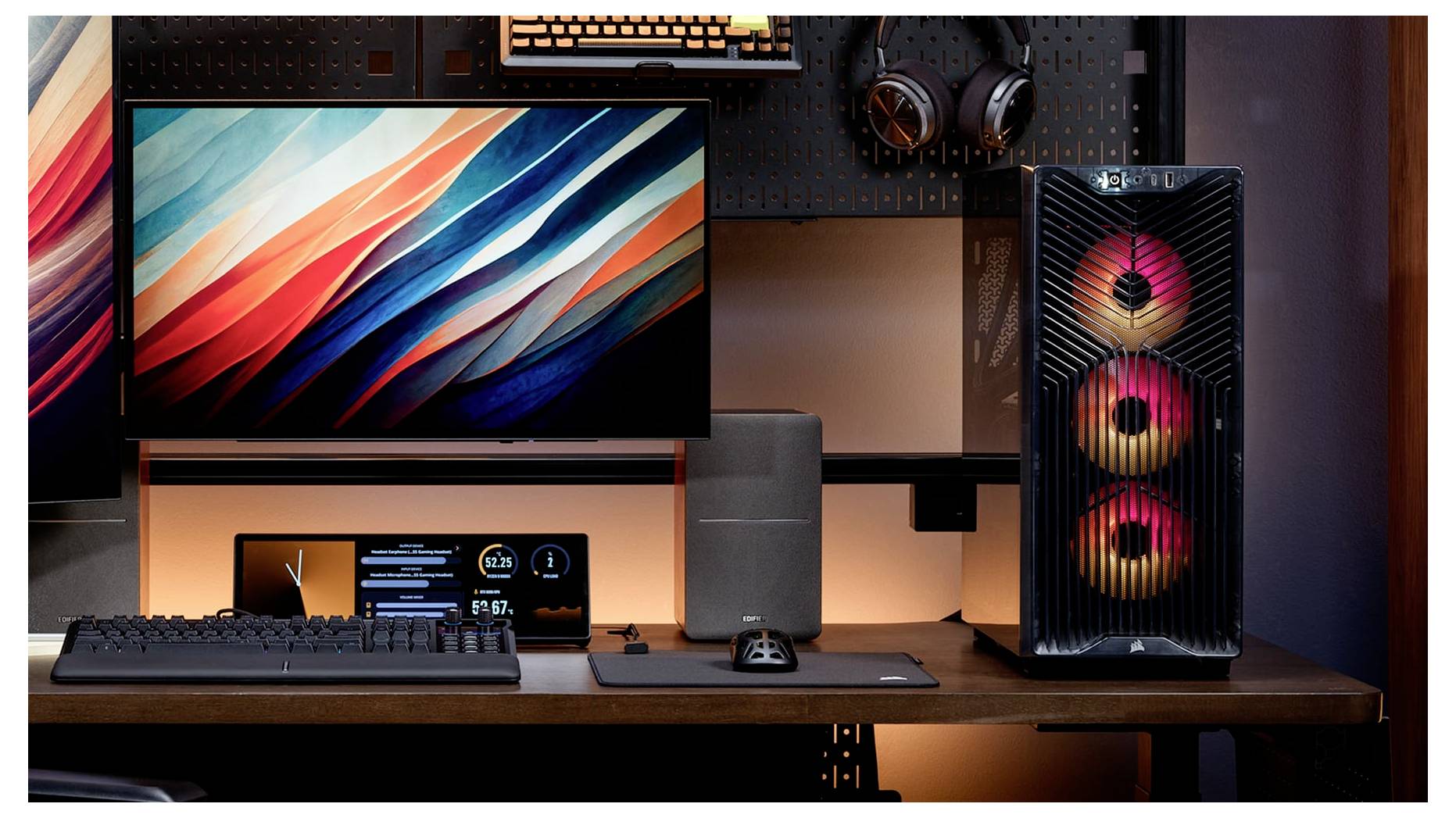 Herní desktopové nastavení s monitorem, klávesnicí, myší, sluchátky a osvětlenou věží PC na dřevěném stole.