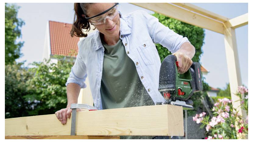 Žena v bezpečnostních brýlích řeže dřevěný trám kruhovou pilou venku v slunečném počasí na stavebním projektu.