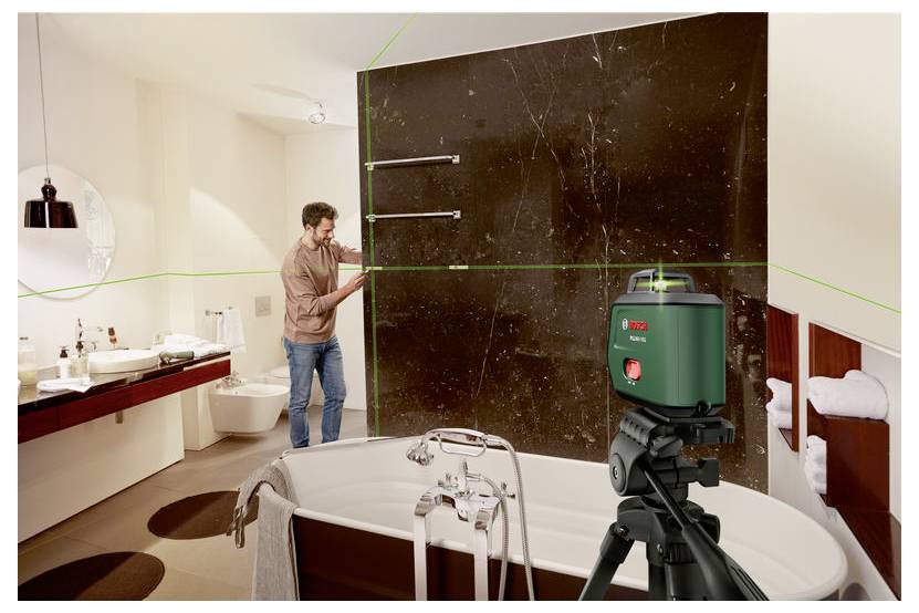 Koupelna ve výstavbě s mužem upravujícím velký tmavý nástěnný panel vedle vany a zeleným laserovým nivelačním přístrojem připevněným na stativu.