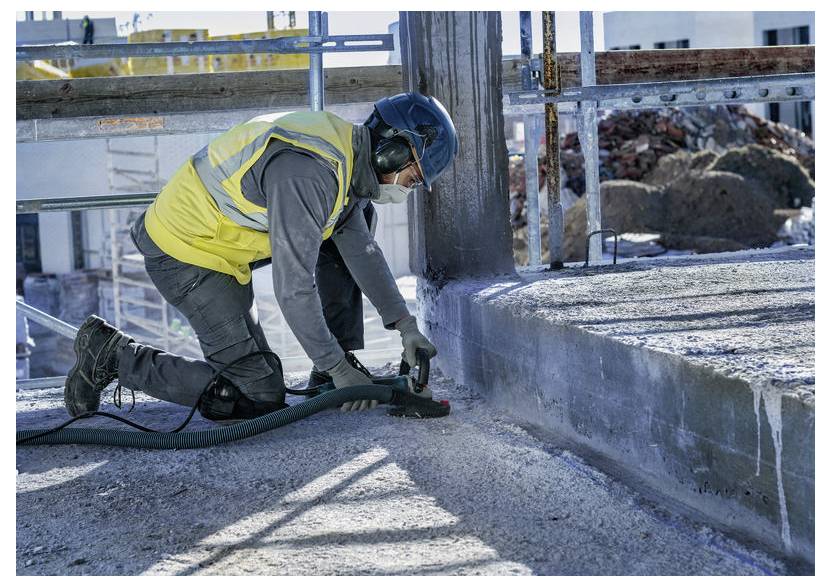 Stavbyní pracovník klečící na betonu, který používá elektrický nástroj k řezání nebo broušení podél hrany desky v exteriéru staveniště.