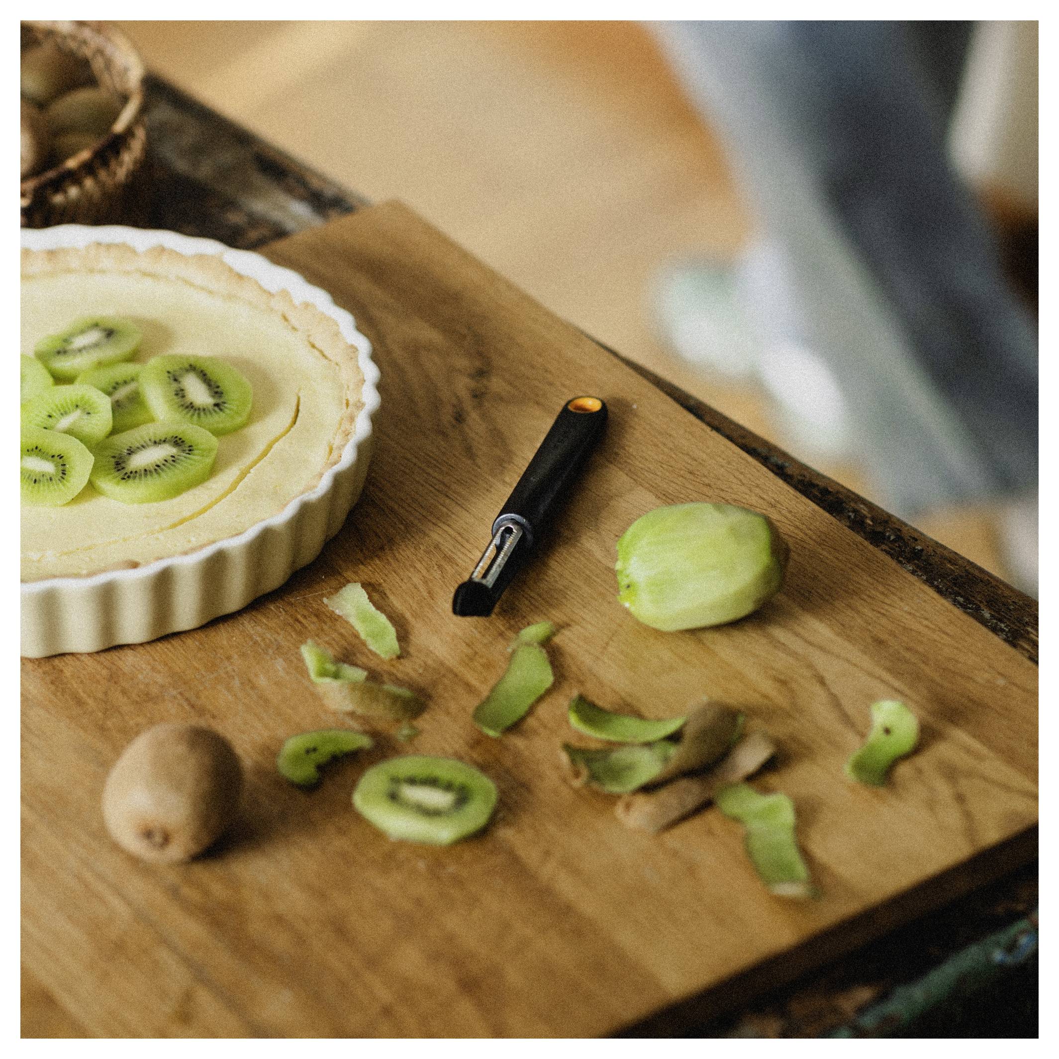 Koláč s kiwi na dřevěné kuchyňské desce se slupovaným a nakrájeným kiwi, slupkami z kiwi a malým loupacím nožem poblíž.