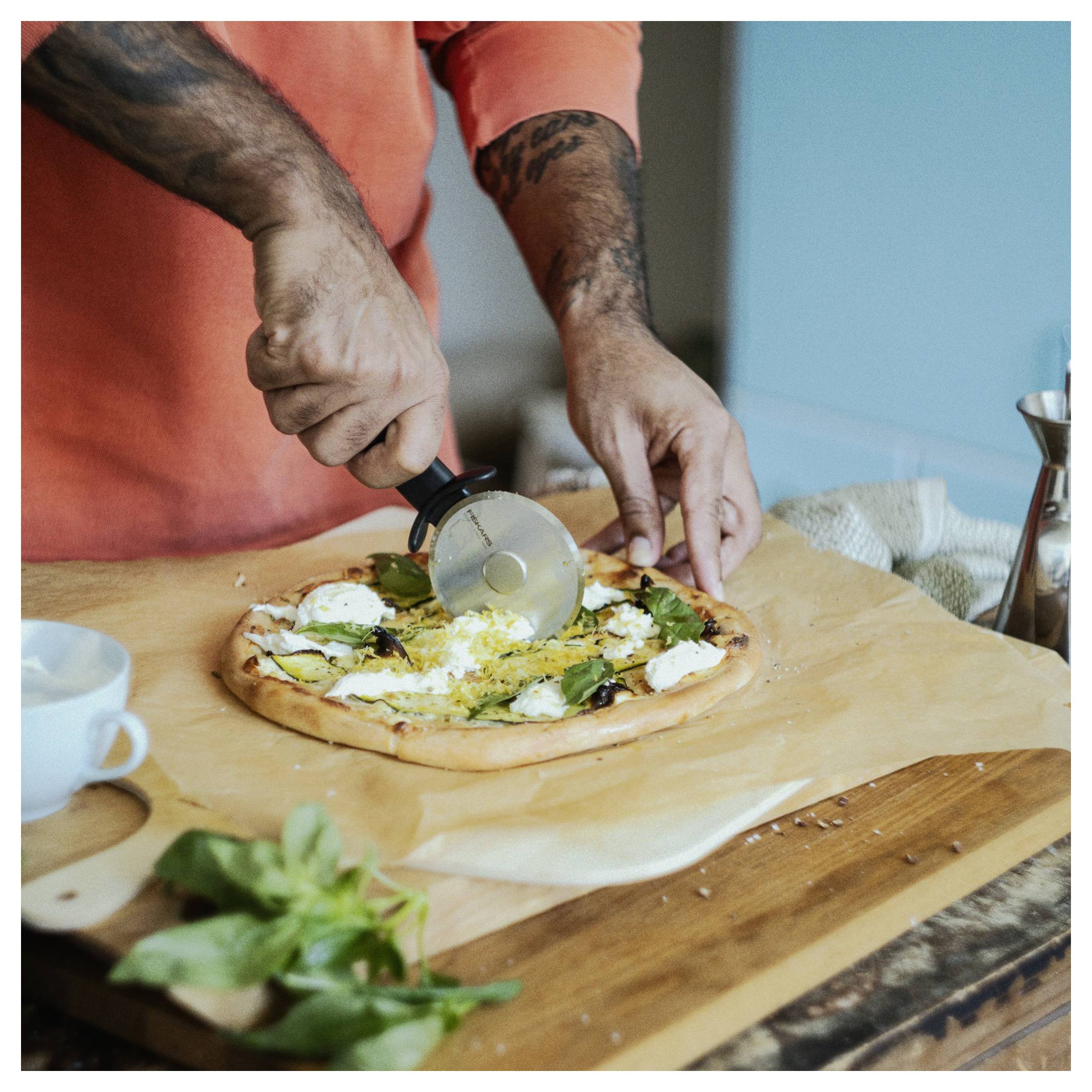 Osoba v oranžovém tričku krájí čerstvou pizzu pomocí krájače na pizzu na dřevěné desce, v blízkosti jsou bazalka a šálek.