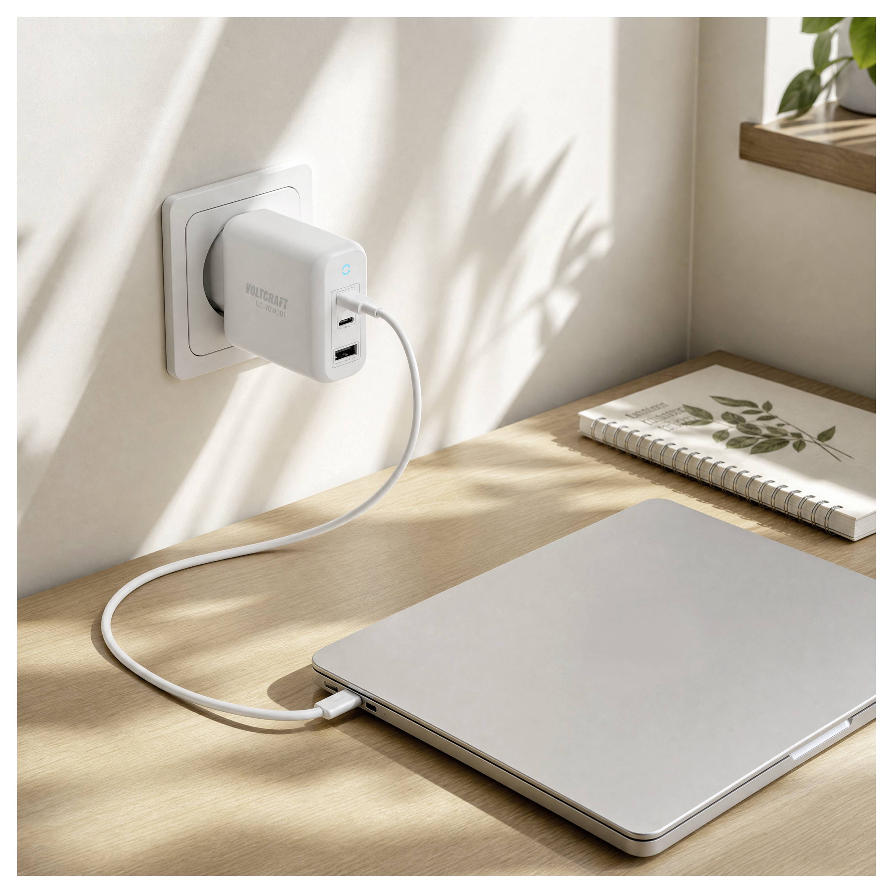 Laptop nabíjející se z elektrické zásuvky přes bílou USB nabíječku na dřevěném stole, s poznámkovým blokem v blízkosti.