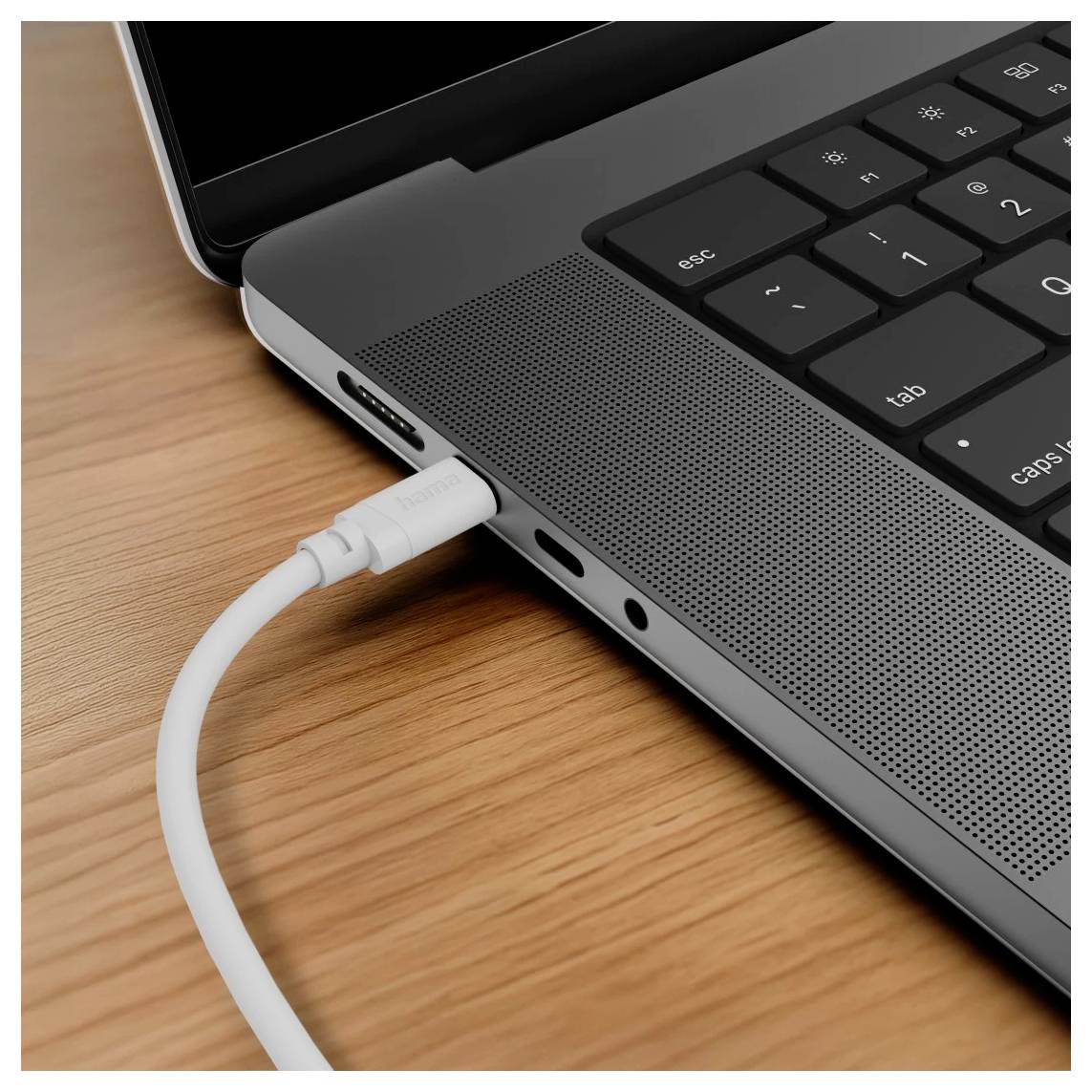 Detailní záběr na hranu laptopa s bílým kabelem zapojeným do bočního portu na dřevěném stole, ukazující nabíjení nebo datové připojení.