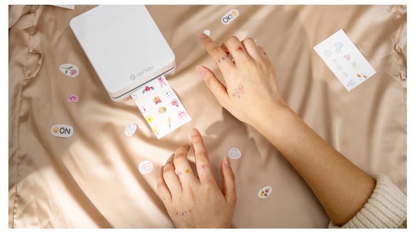 Dvě ruce používají malou bílou fotoprinter Canon k tisku barevného listu samolepek na posteli, kolem něhož jsou rozptýleny dekorativní samolepky.