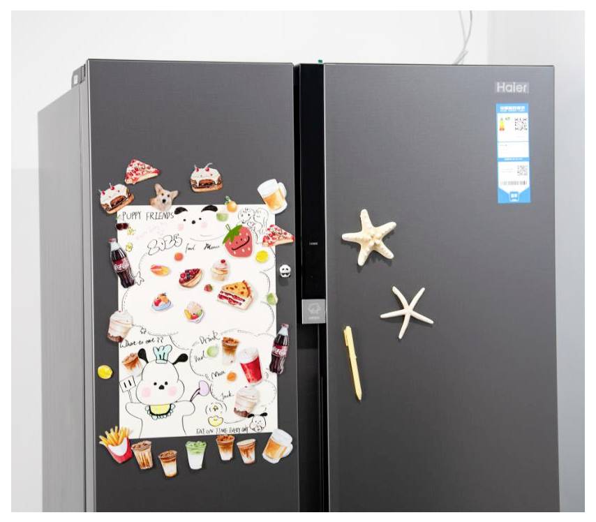 Dvoudvéřová černá lednice Haier s dětskými kresbami a magnetky na zákusky na levých dveřích, hvězdice a magnetka ve tvaru tyčinky na pravých dveřích.