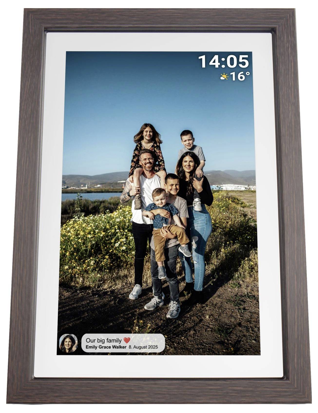 Digitální fotorámeček zobrazující rodinný portrét venku; překryv na obrazovce ukazuje čas 14:05, 16 stupňů a titulek "Naše velká rodina" se jmény a datem srpen 2025.