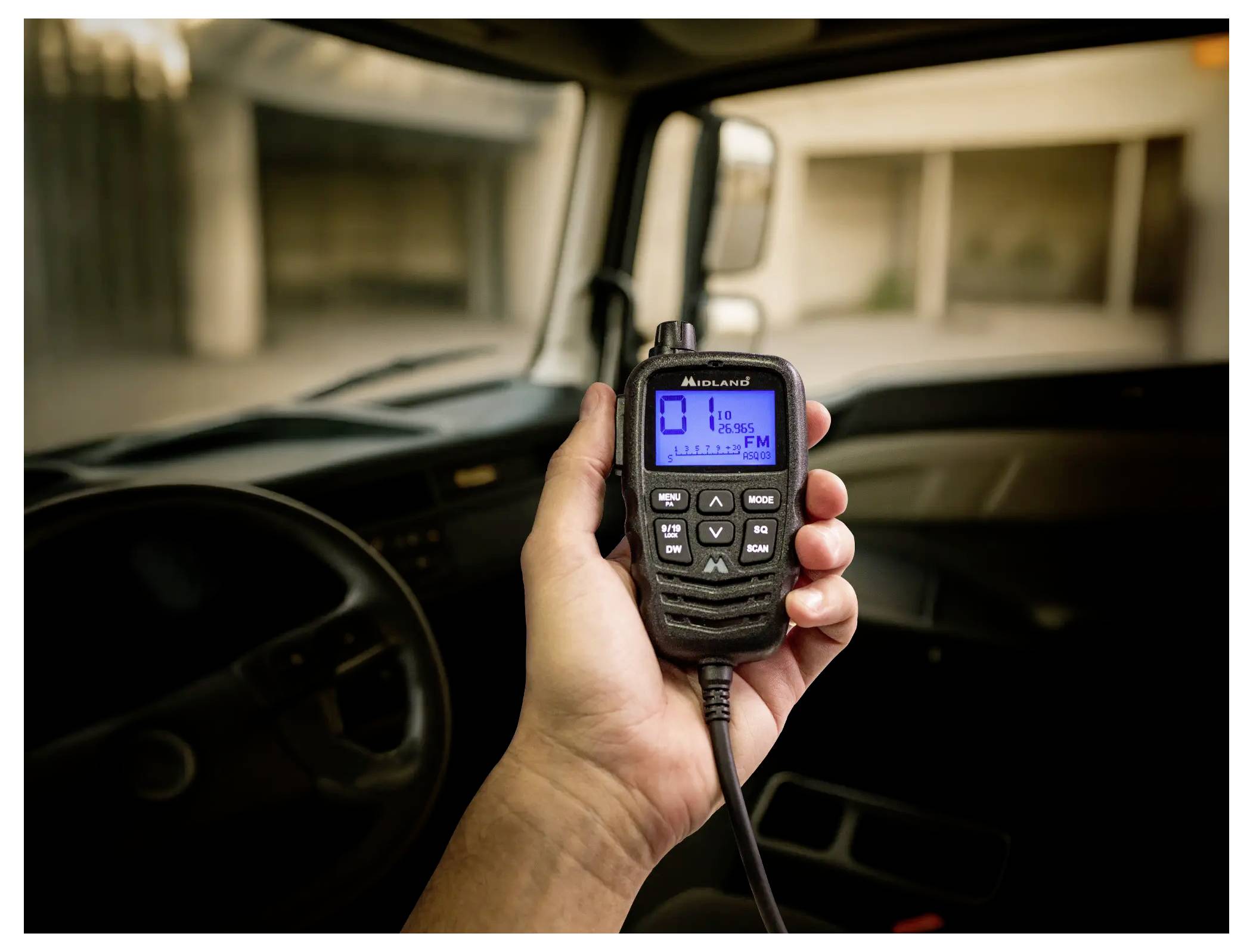Ruka držící radiostanici Midland do dvou směrů uvnitř automobilu s modrým zapnutým displejem a připojeným krčeným kabelem.
