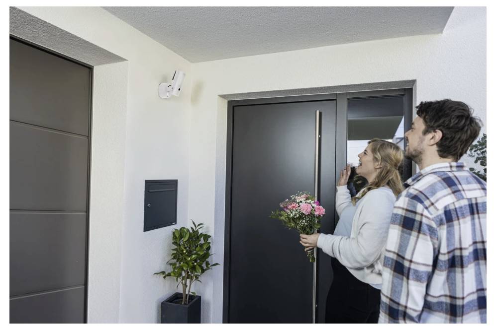 Pár stojí u dveří moderního bytu, žena drží růžové květiny a zvoní na zvonek.