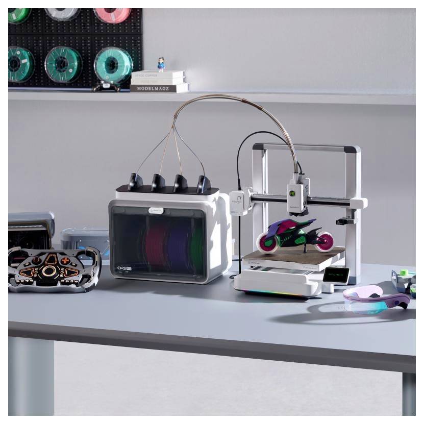 Tiskárna 3D na stole tiskne malý barevný model, kolem ní jsou uspořádány prototypové díly a součástky tiskárny.