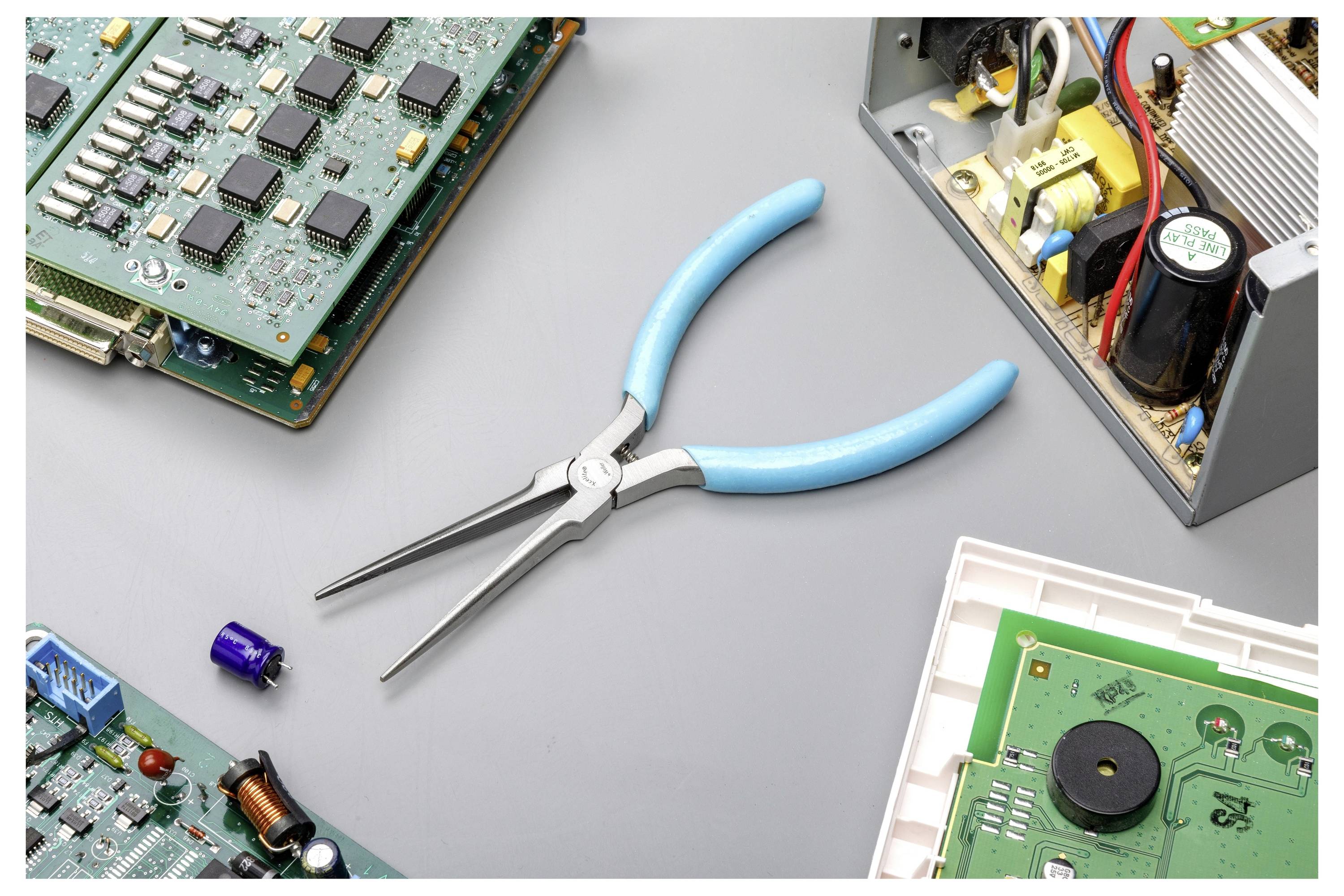Modré kleště soustředěné mezi různorodými elektronickými deskami plošných spojů a součástkami na šedé pracovní ploše.