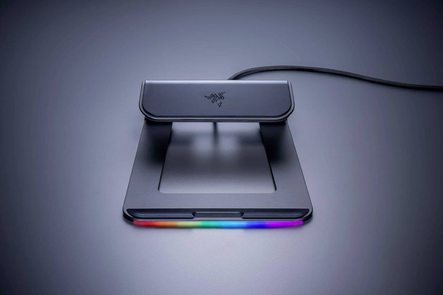 Černý stojan na notebook s duhovým LED páskem podél přední hrany, umístěný na tmavém povrchu s kabelem vycházejícím ze zadní strany.