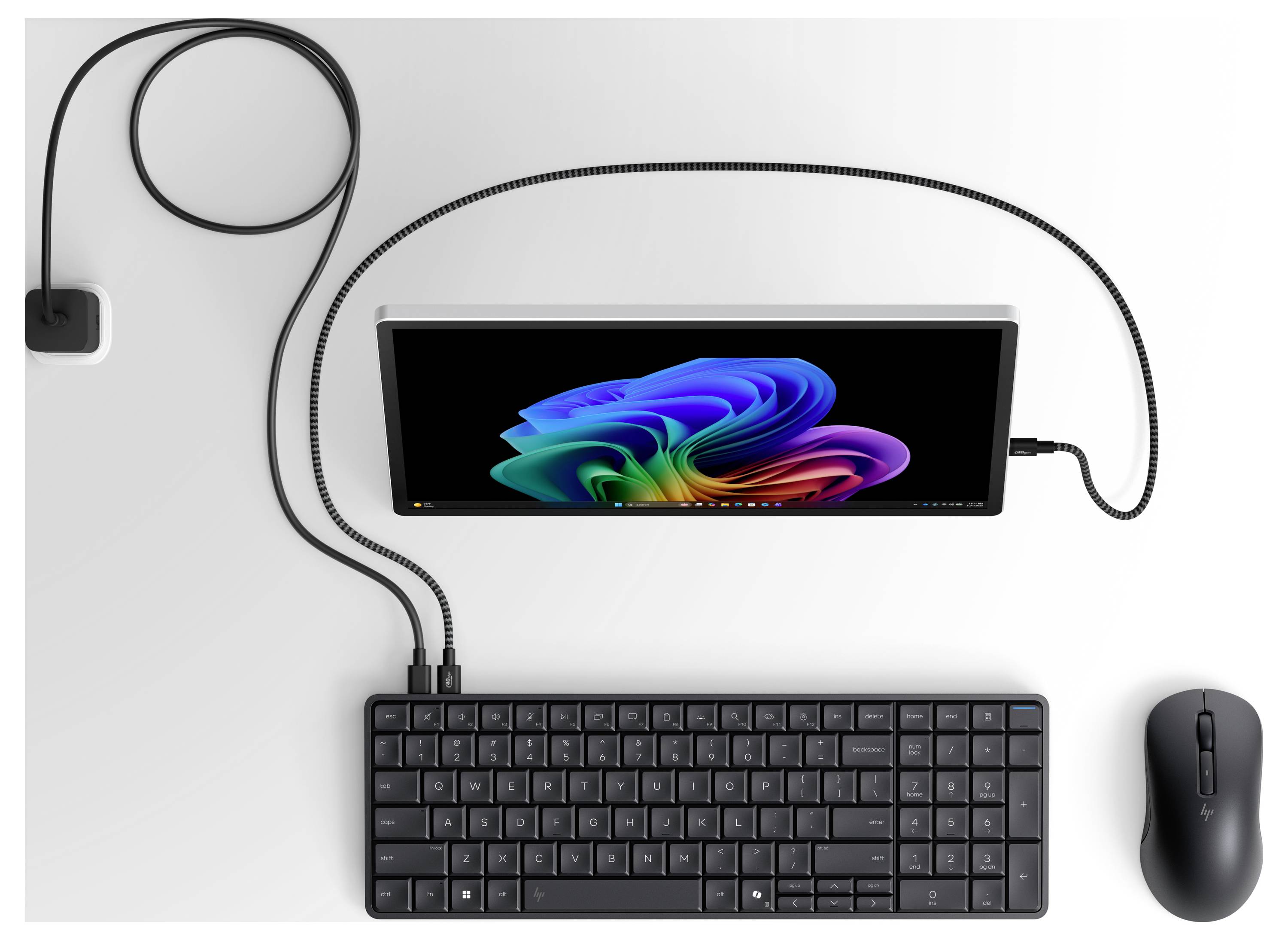 Pohled shora na drátěné stolní zařízení se středovým monitorem, klávesnicí, myší a napájecím adaptérem spojenými kabely na bílém povrchu.