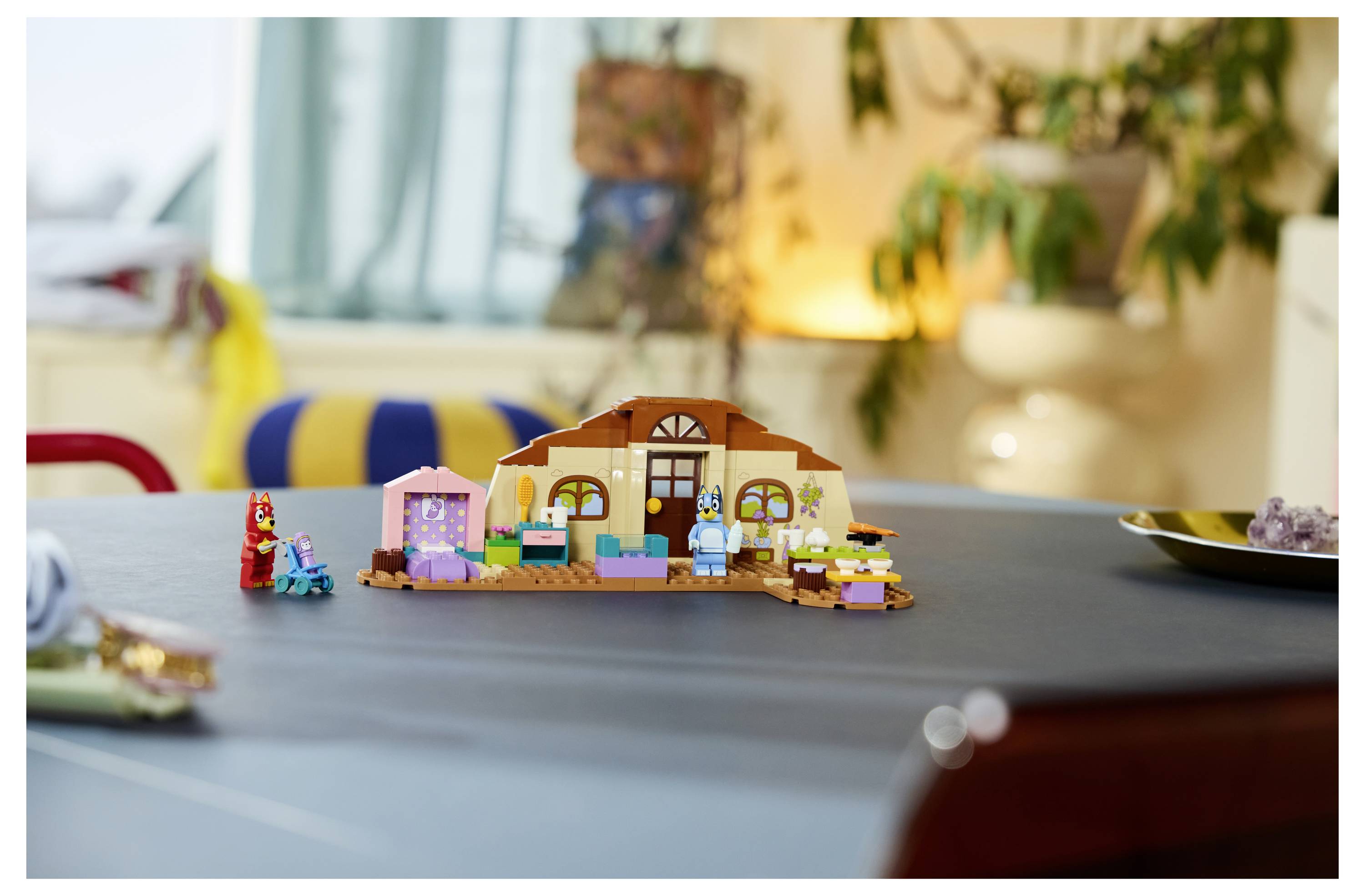 Malá LEGO stylem postavená scéna domu na stole s malými figurkami a doplňky v útulném rozmazaném prostředí obývacího pokoje.