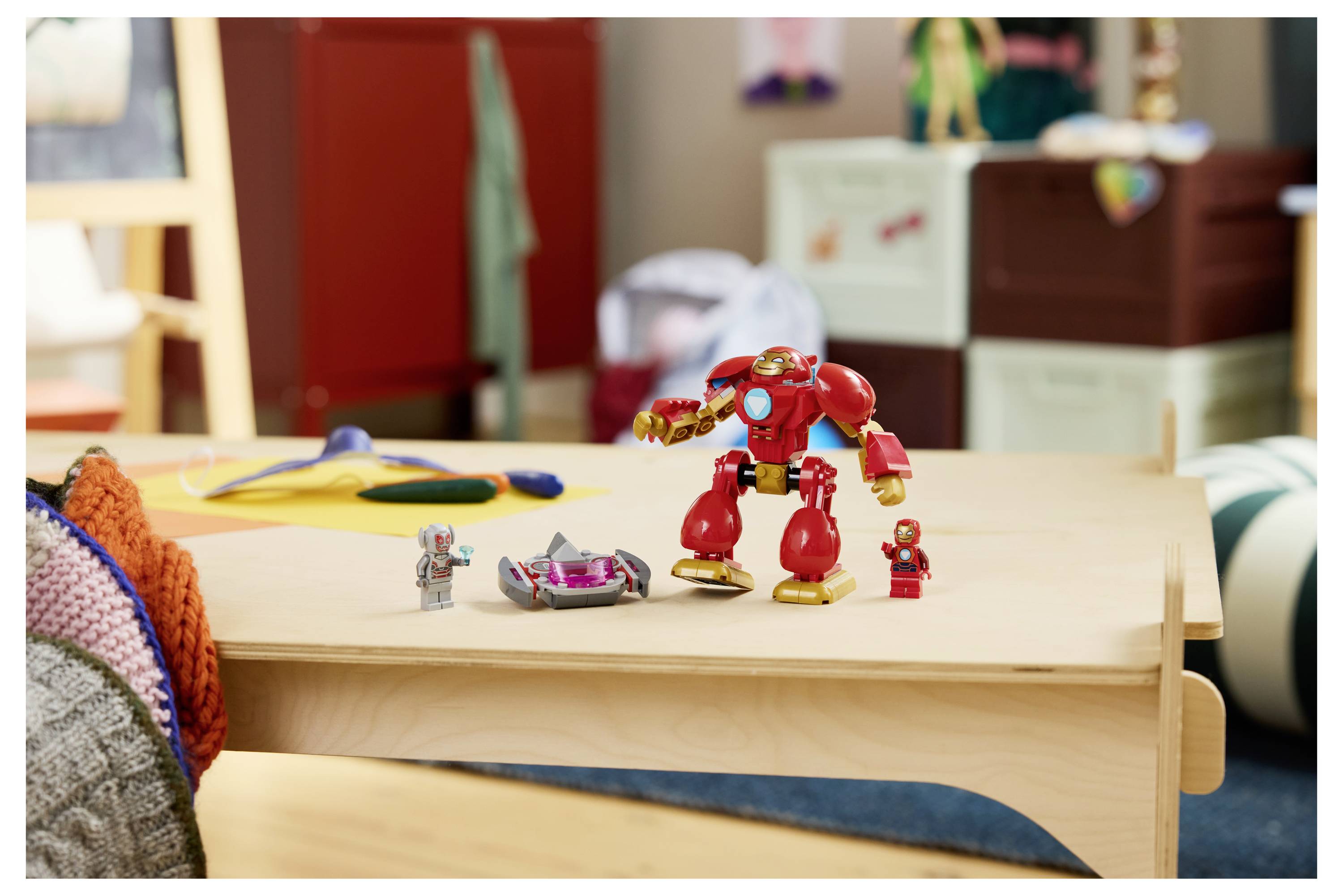 Červený hračkový robot stojí na nízké dřevěné tabulce se dvěma malými hračkovými figurkami a vozidlem vedle něj v dětském pokoji.