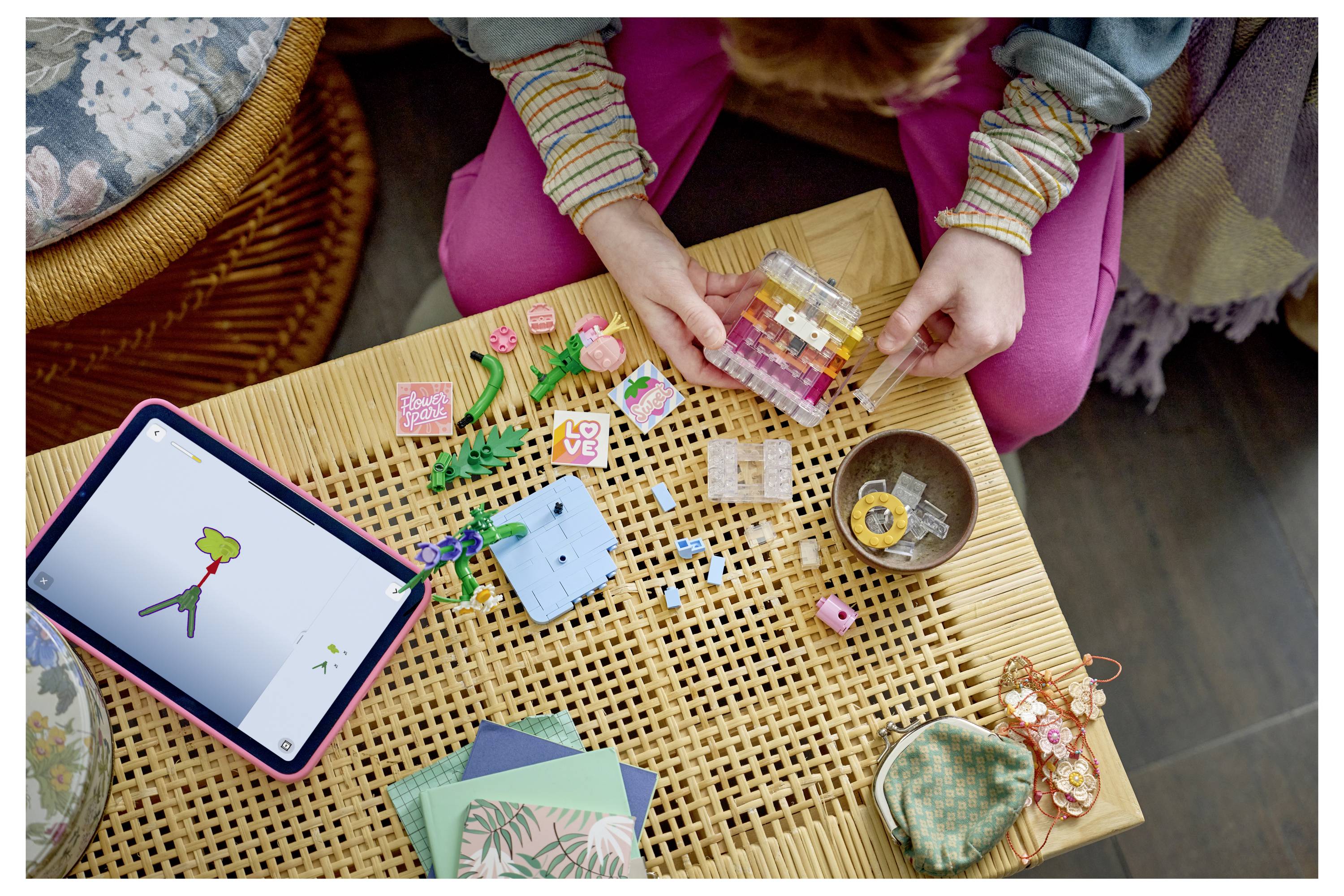 Dva lidé skládají barevné LEGO kostky na tkané stole vedle tabletu zobrazujícího digitální kresbu, s výtvarnými materiály a malými kontejnery kolem nich.