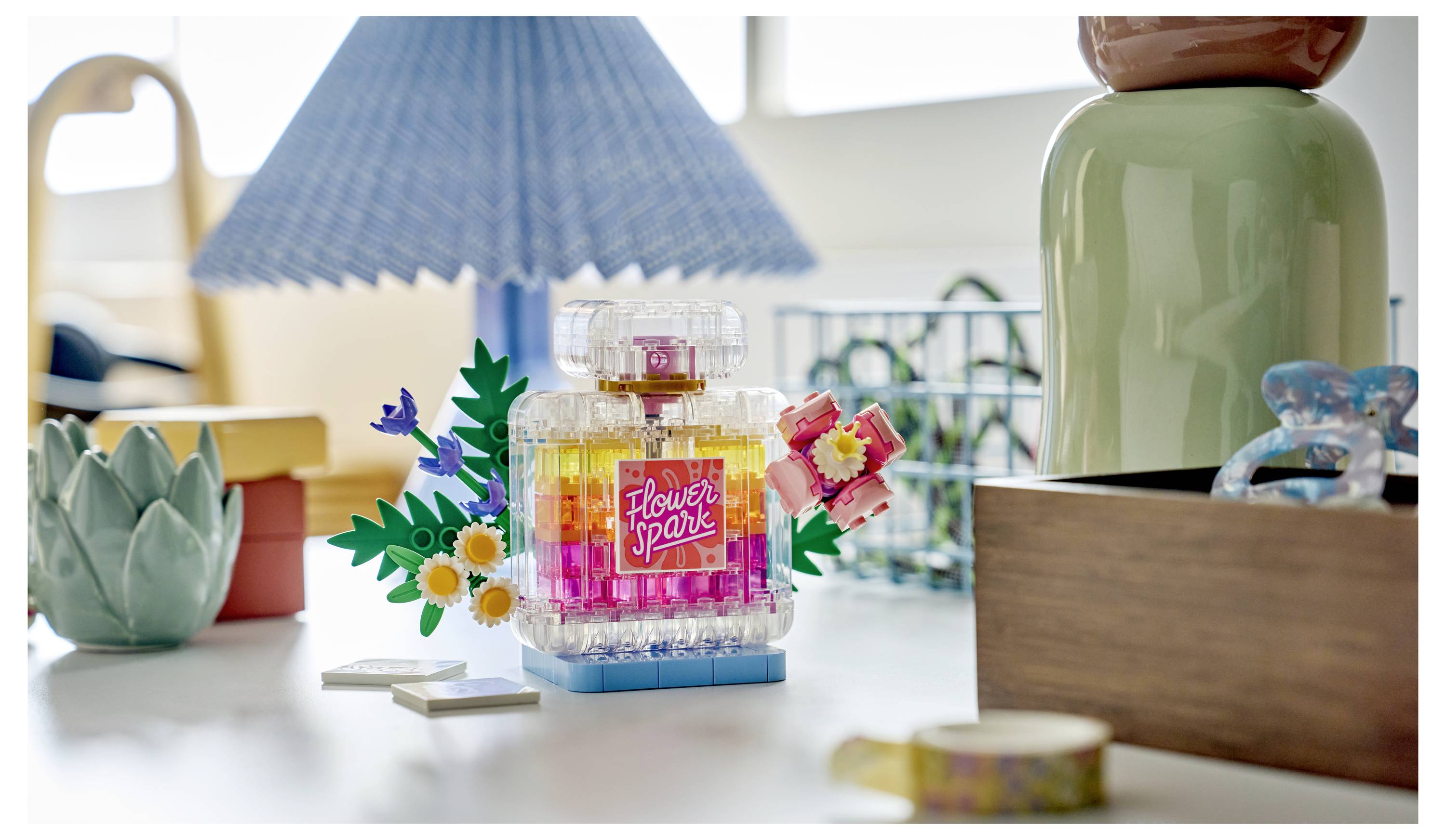 Barevný LEGO-style model láhve s parfémem s názvem "Flower Spark" na desce stolu, obklopen hravou dekorací a modrým stínidlem lampy.