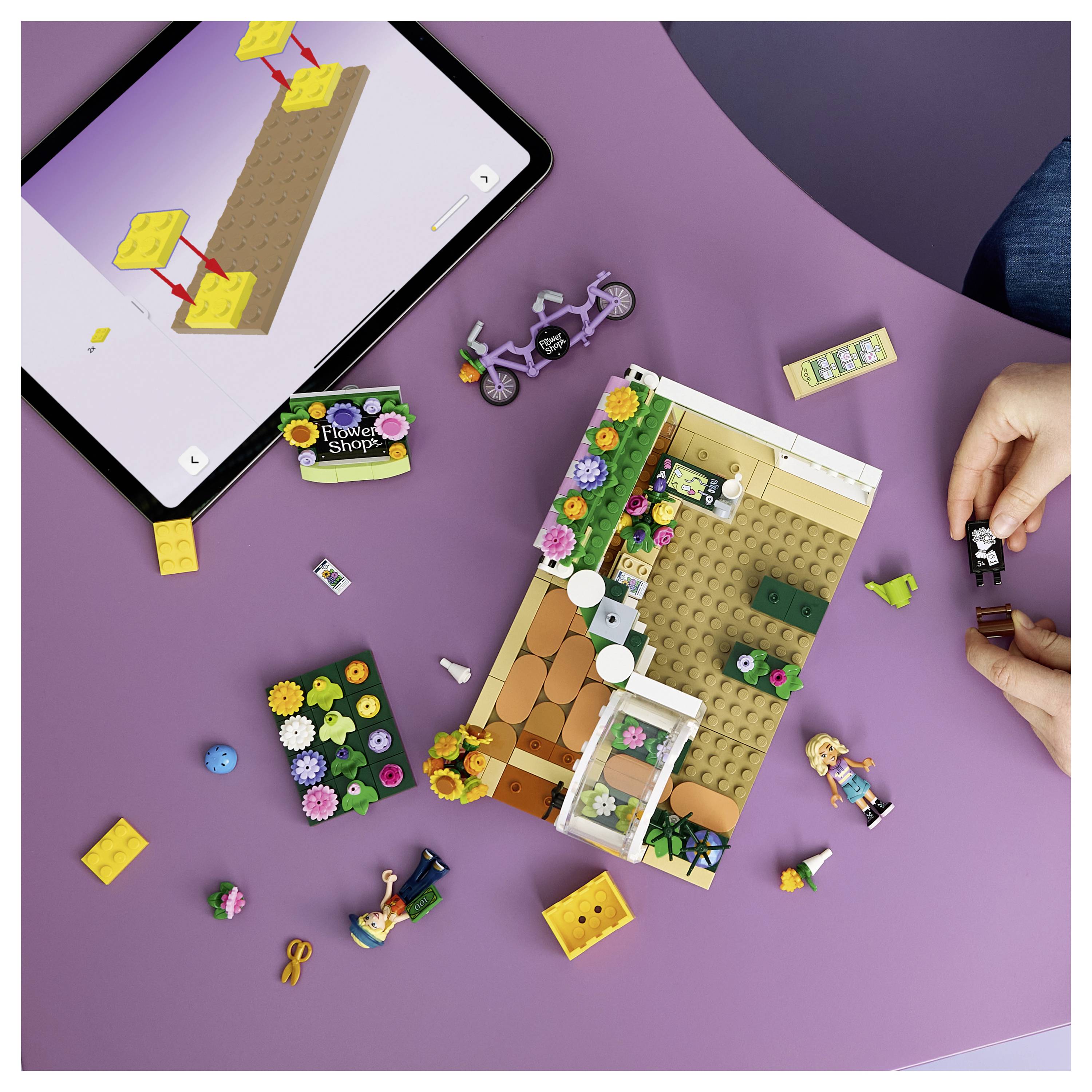 Ruce skládající stavebnici květinářství ve stylu LEGO vedle tabletu zobrazujícího pokyny pro stavbu, s volnými dílky a jízdním kolem na fialkovém povrchu.