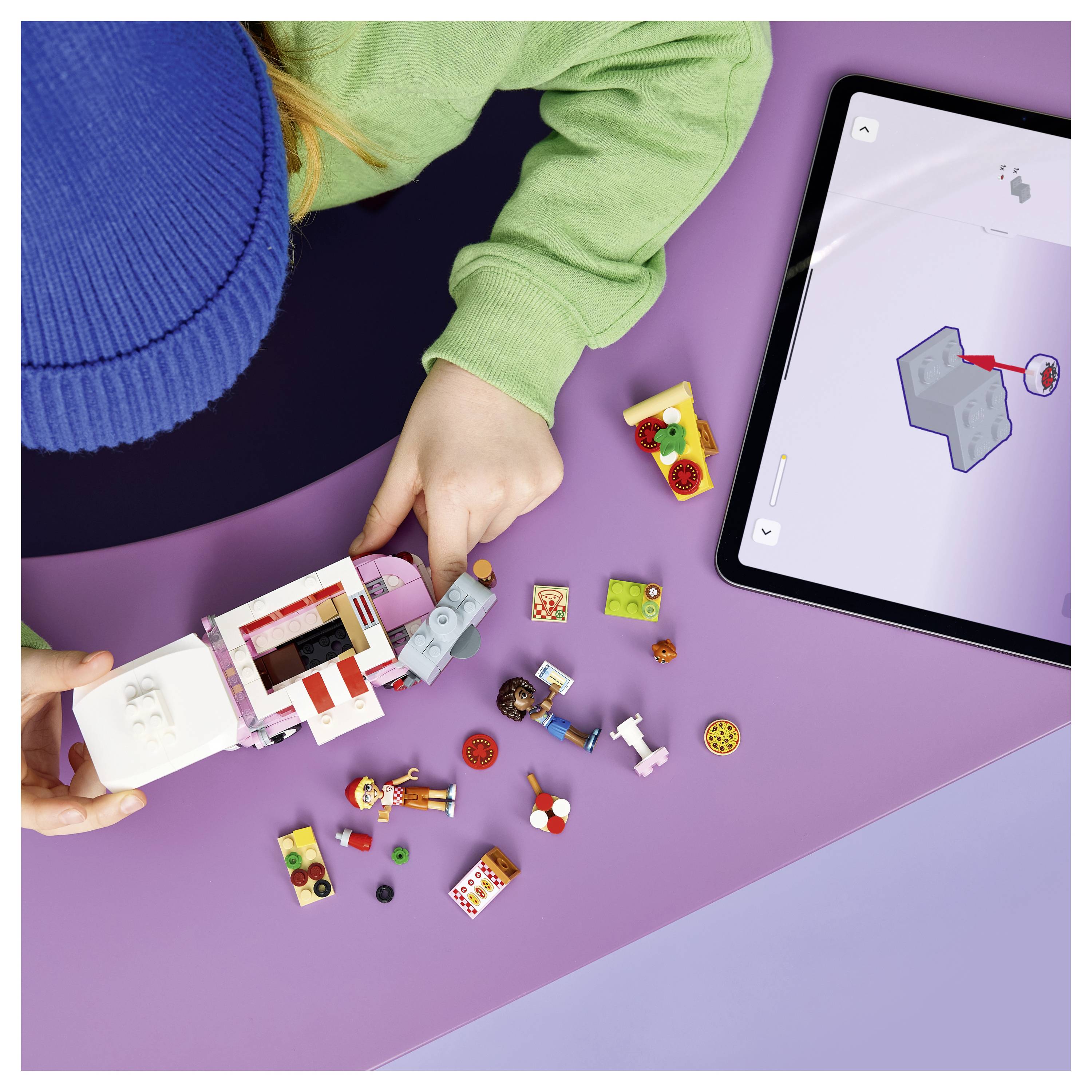 Dítě staví model LEGO podle papírových pokynů, zatímco tablet zobrazuje odpovídající 3D návod pro montáž na fialovém stole.