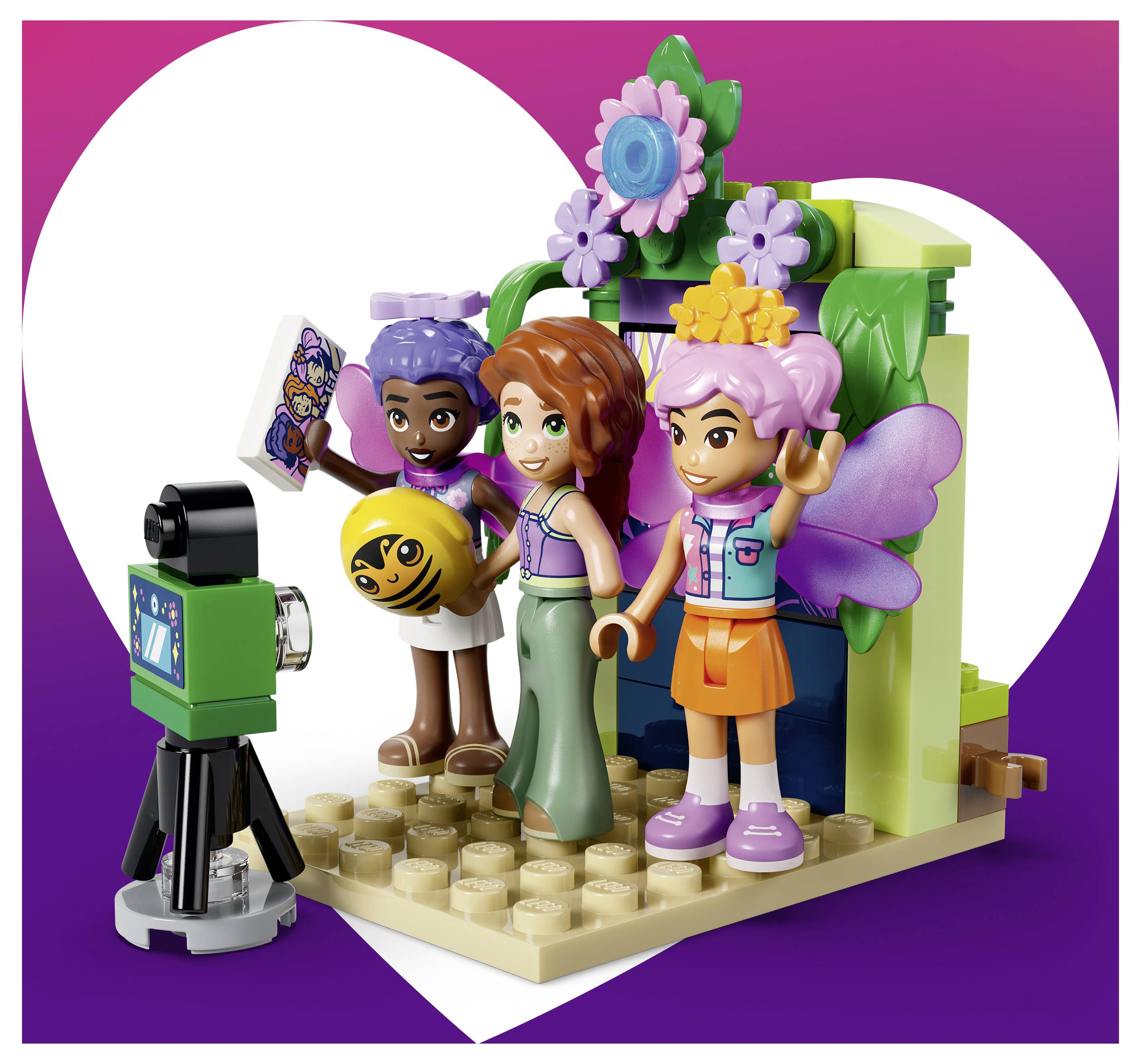 Tři LEGO stylizovaní přátelé pózují na platformě s fotoaparátem na stativu, drží ceduli a míč, před pozadím v podobě srdce.
