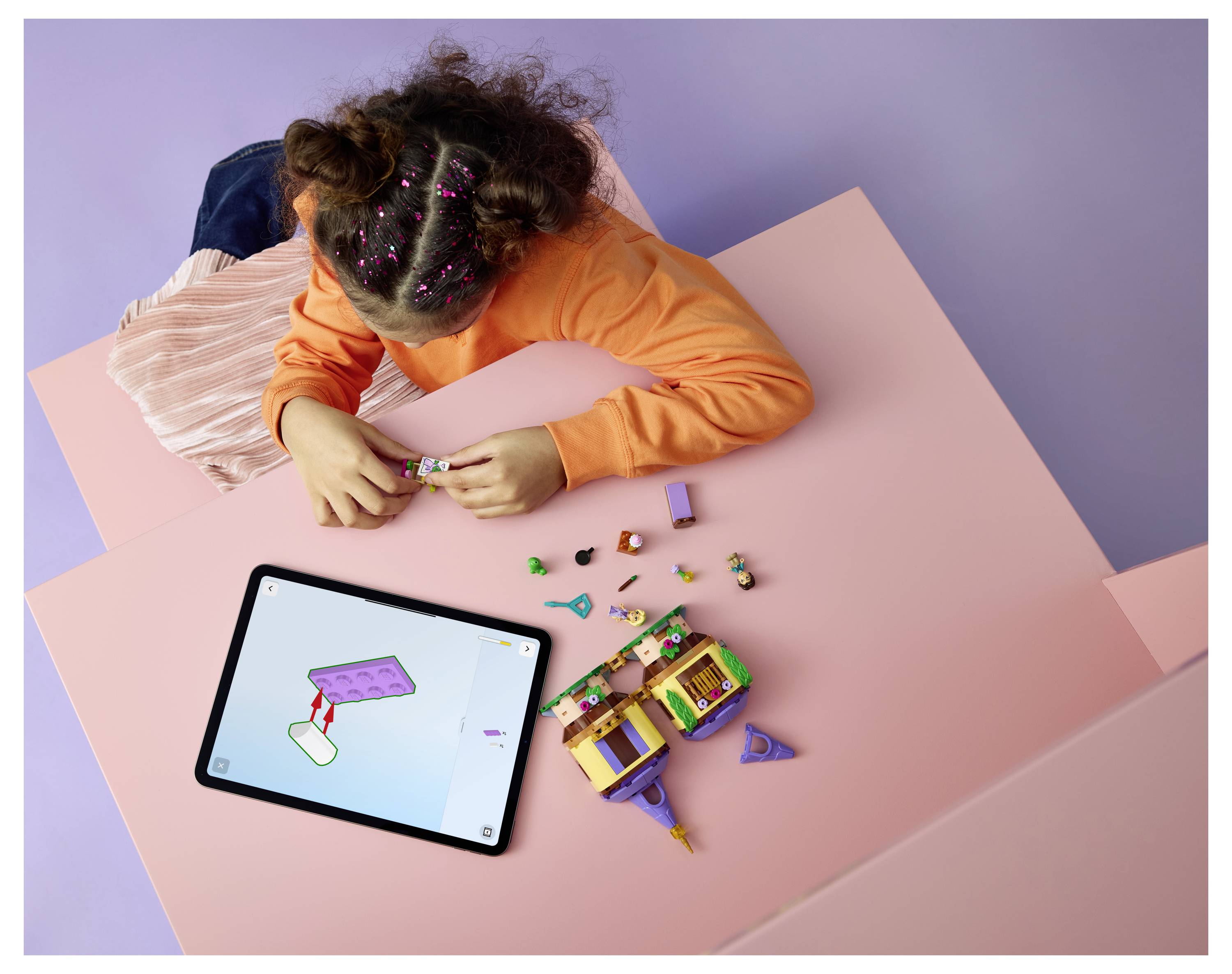 Dítě buduje malou barevnou sadu hraček na růžovém stole a používá tablet jako průvodce.