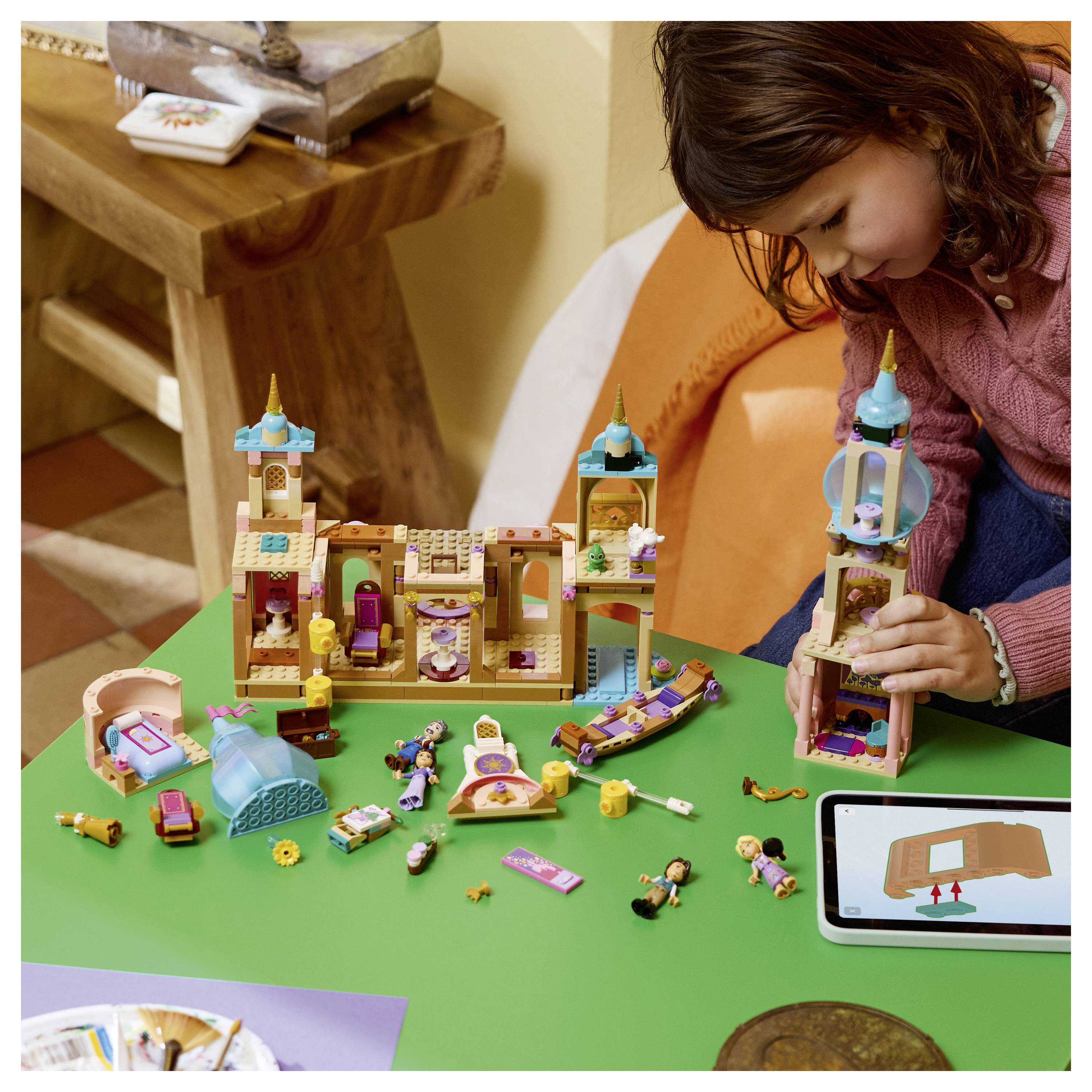 Mladá dívka staví barevný hrad ve stylu LEGO na zeleném stole, zatímco tablet zobrazuje stejnou sadu jako návod k montáži.