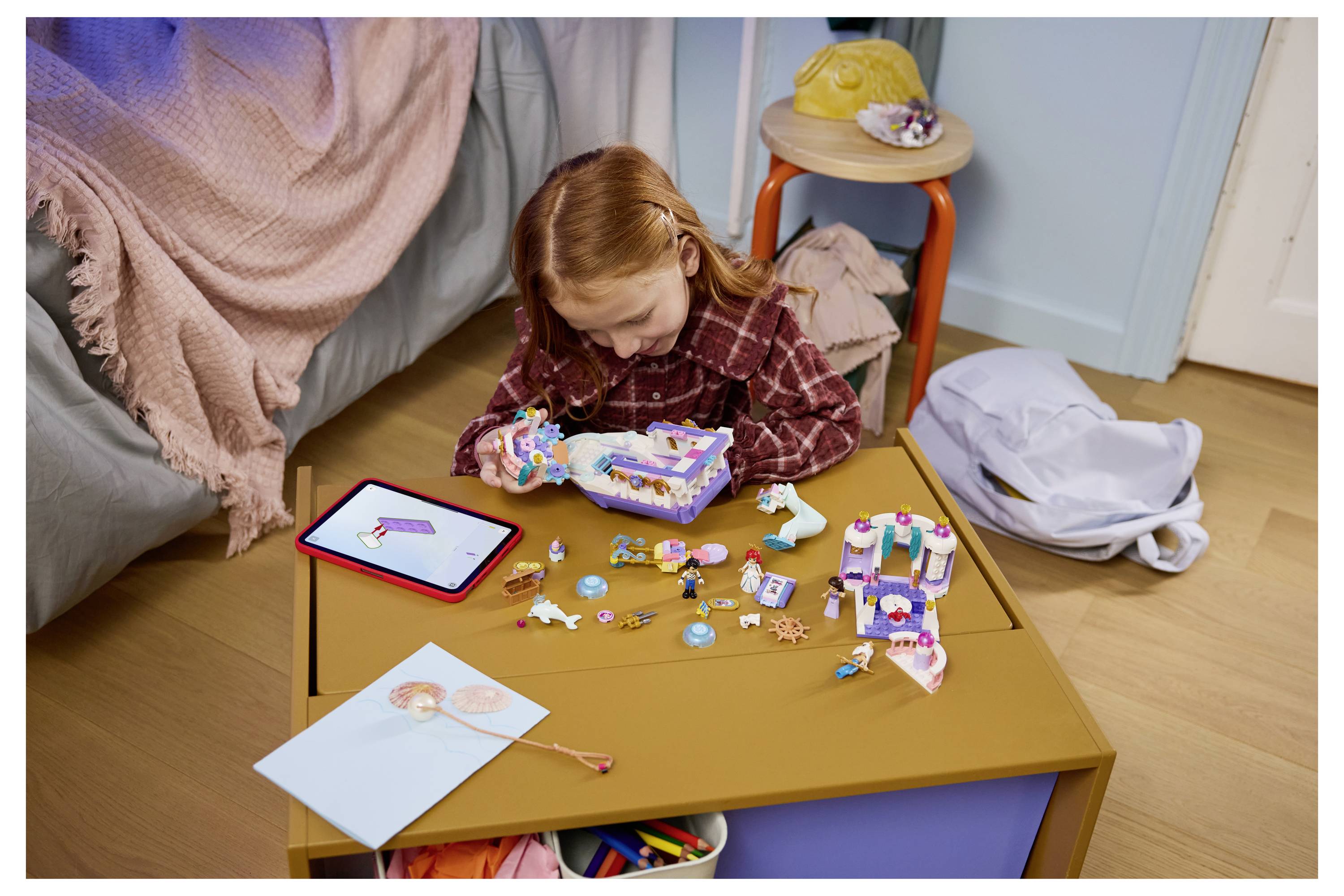 Malá dívka sedí na podlaze a skládá barevnou sadu hraček, obklopená rozptýlenými díly, tabletem a tvůrčími materiály na malém stole.
