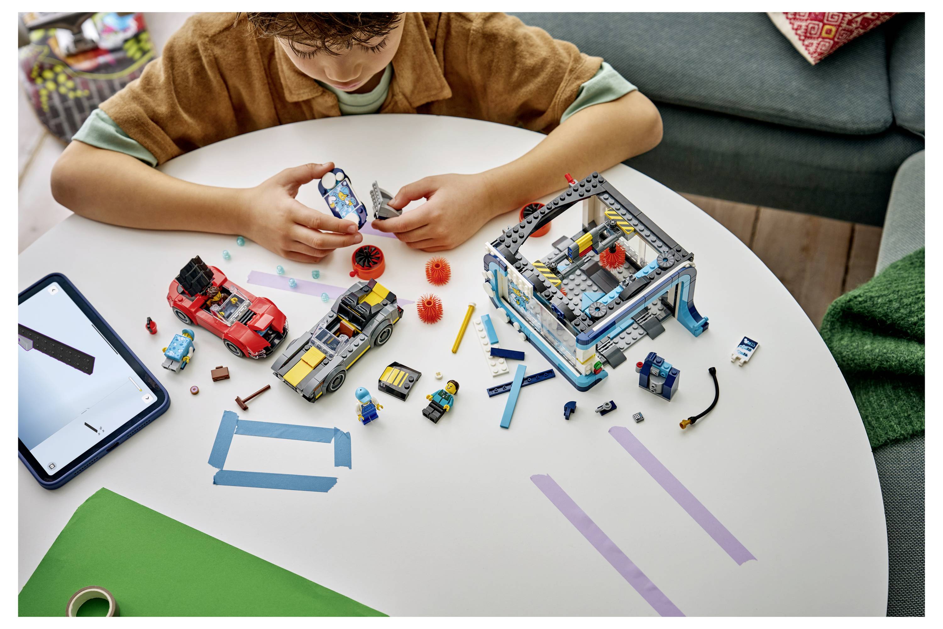 Dítě skládající model ve stylu LEGO na stole s rozptýlenými kostkami, instrukcemi na tabletu a částečně postavenými hračkami a stavbami vozidel.