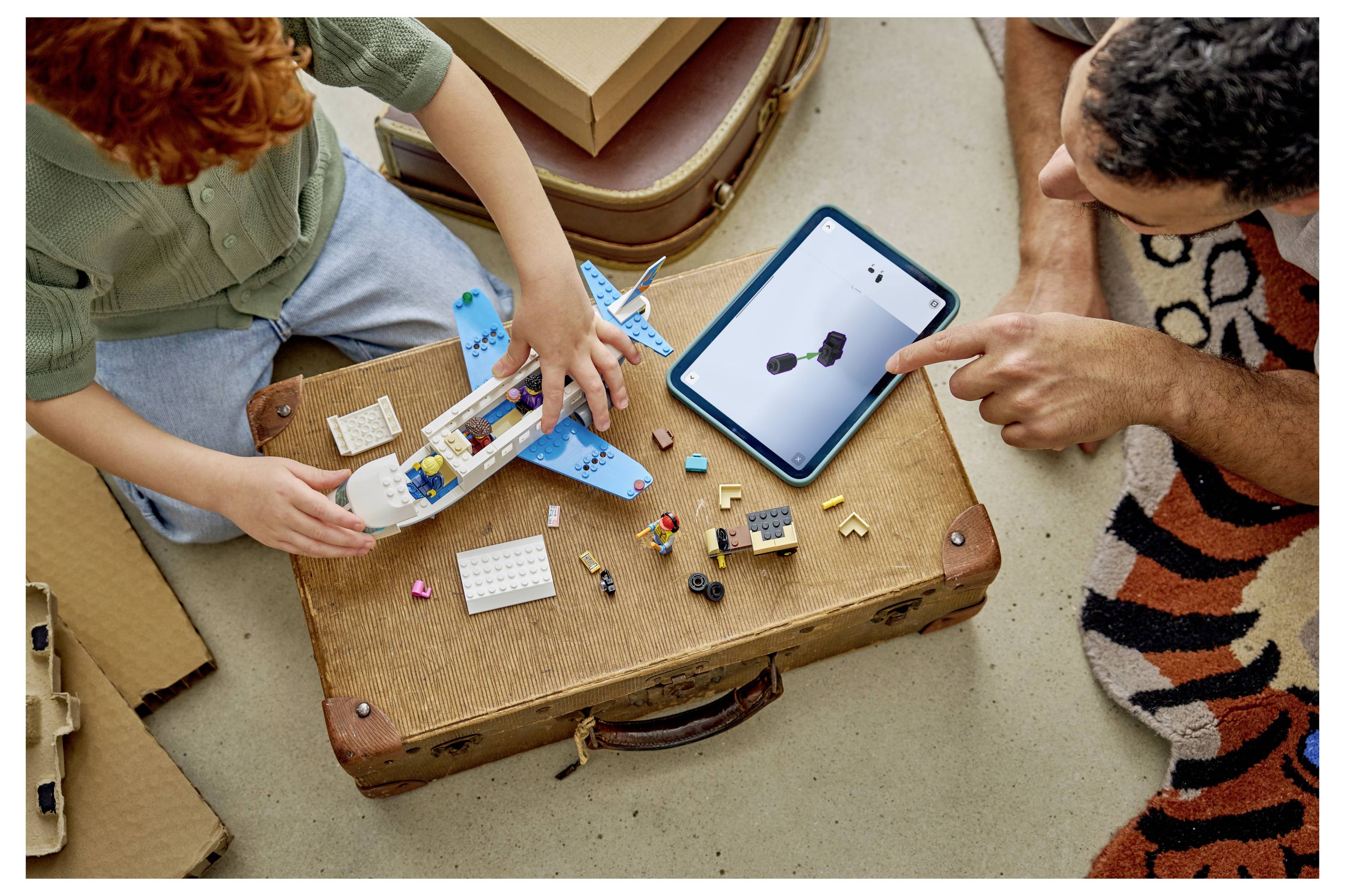 Dva lidé a dítě staví malé hračkářské letadlo z kostek podobných LEGOu, zatímco další osoba ukazuje na tablet zobrazující 3D model.
