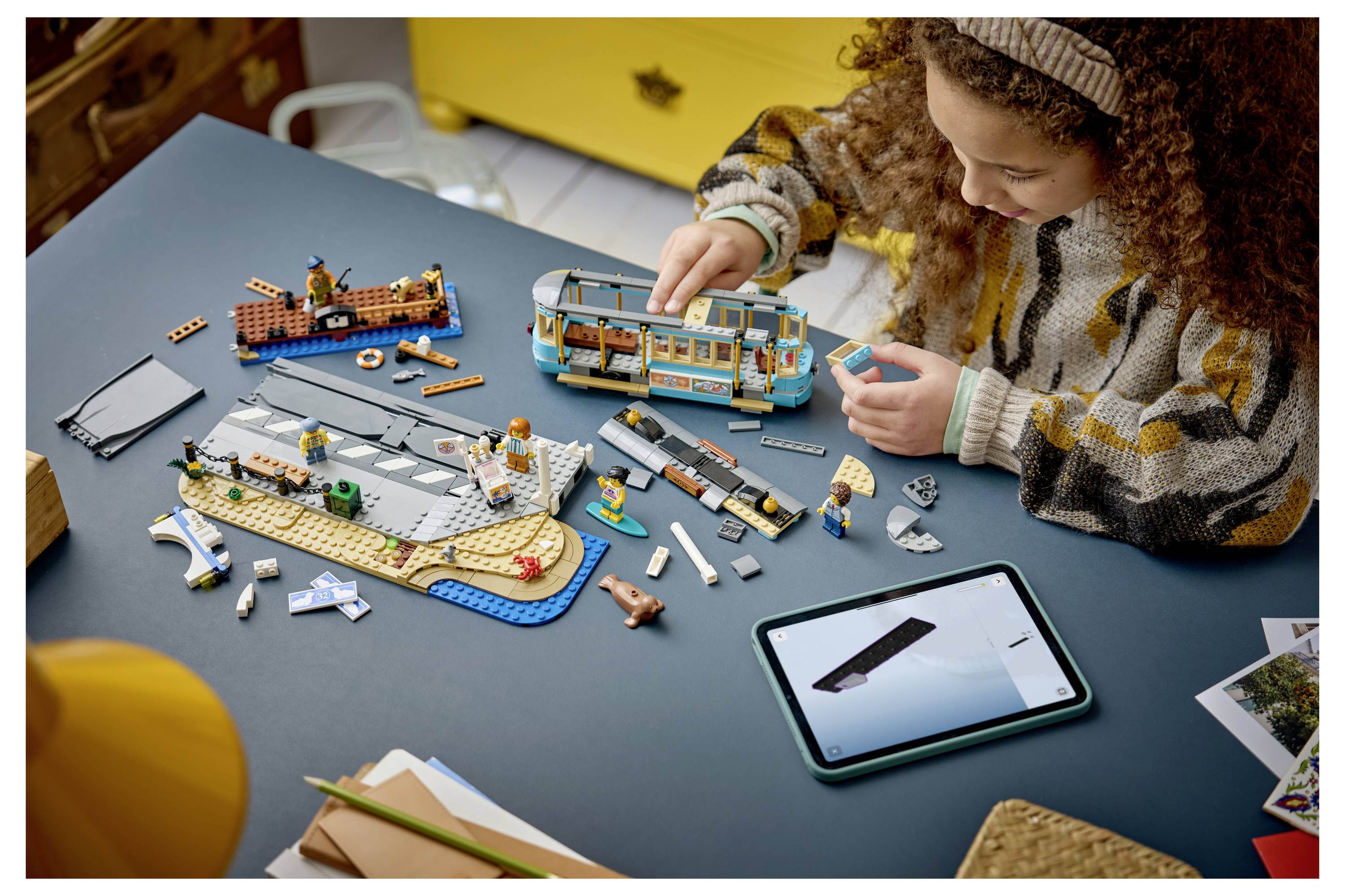 Dítě stavějící model ve stylu LEGO na stole s pokyny na tabletu, obklopené rozptýlenými kusy a jinými sestavenými hračkami.