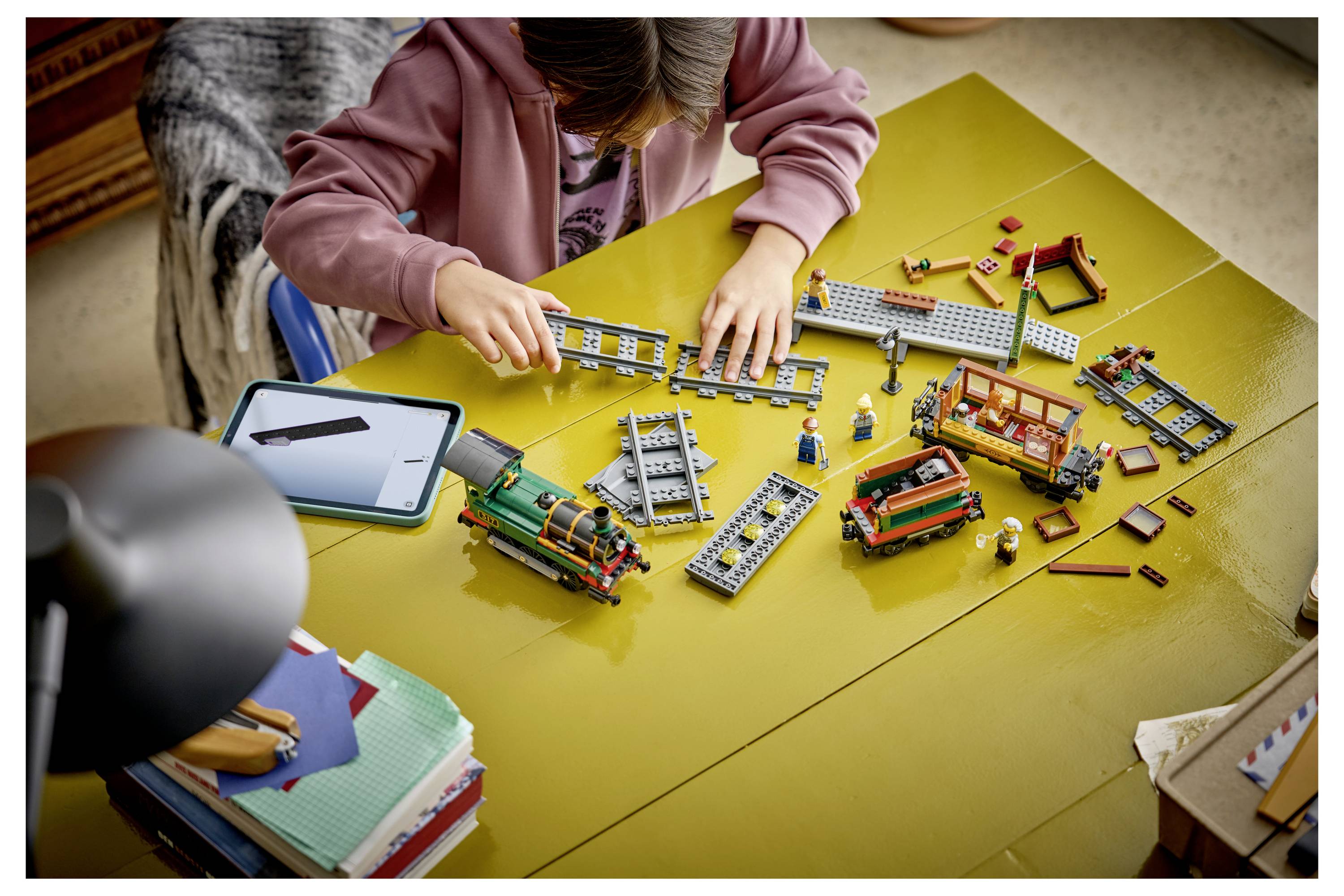 Dítě skládá sadu LEGO vlaku u žlutého stolu, s kolejnicemi, částmi vlaku a malými figurkami rozloženými kolem něj.