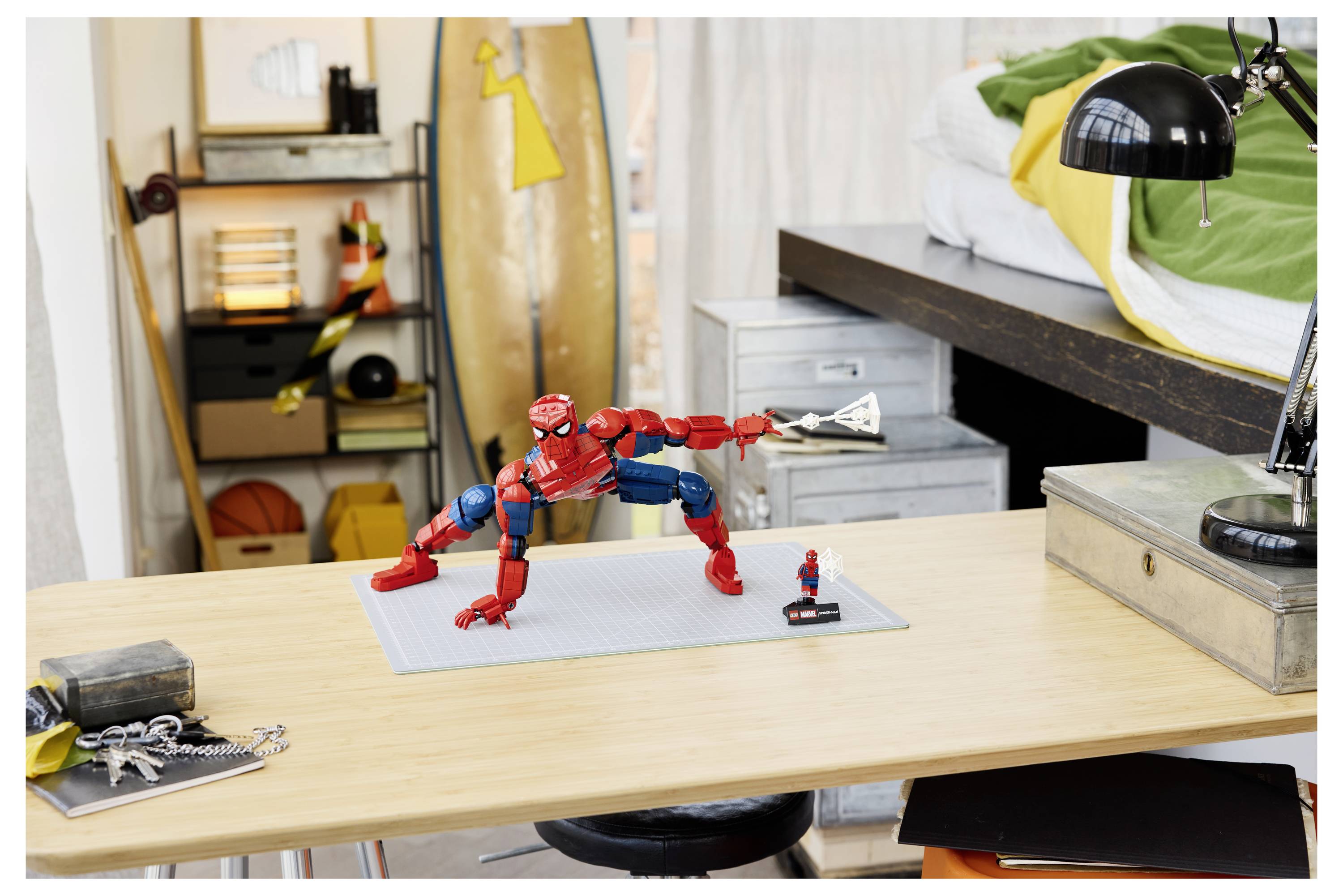 Figurka Spider-Mana v akční póze na stole v ložnici, s malou minifigurkou vedle ní a skateboardem a lampou v pozadí.