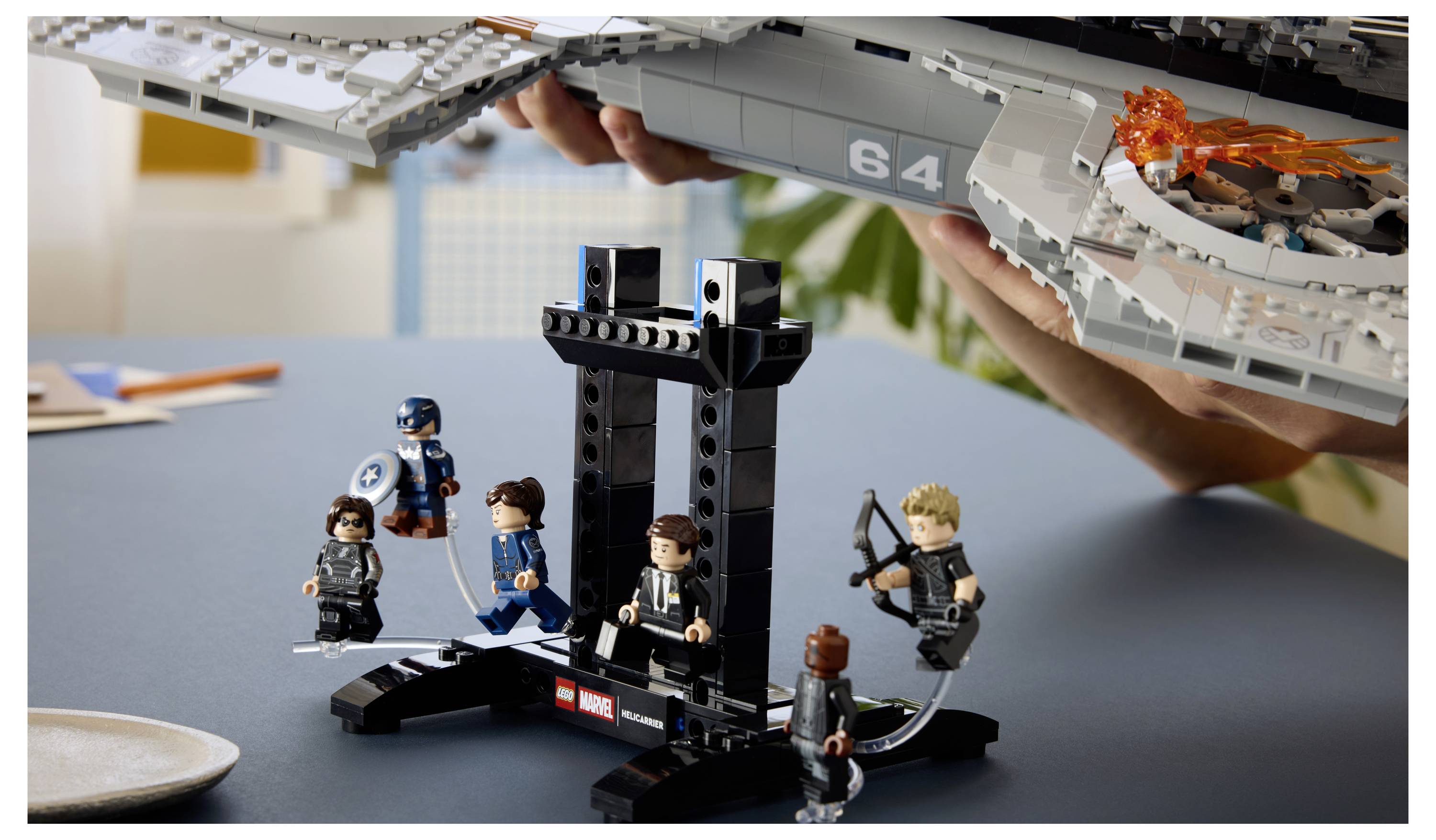 LEGO Marvel display s pěti minifigurkami kolem černého podstavce, přičemž ruce drží velkou šedou loď nad hlavou s malým oranžovým detailem výbuchu na lodi.