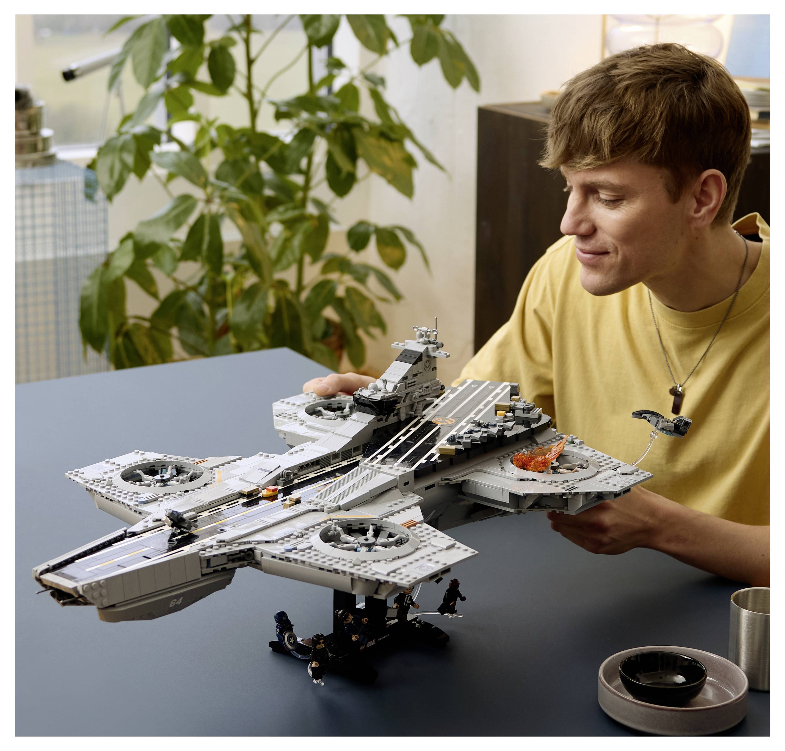 Mladý muž v žlutém tričku skládá na stole velký šedý model letadla nebo vesmírné lodi ve stylu LEGO.